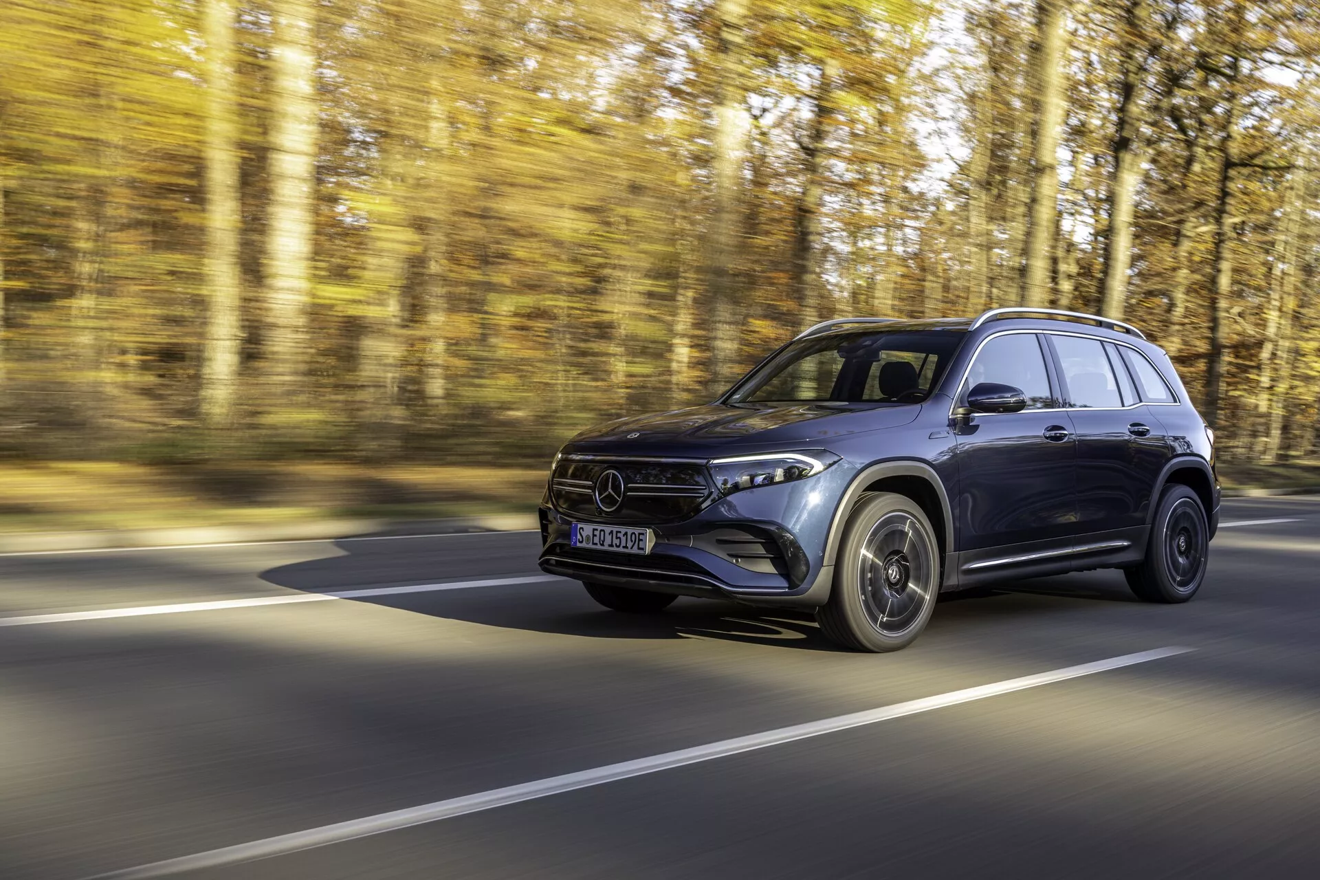 SUV Mercedes EQB 2021 en mouvement sur une route forestière, capturé sous un angle côté avant avec une vue claire du côté conducteur; doté de phares LED, d'une calandre distinctive, de rétroviseurs latéraux avec clignotants intégrés et de roues en alliage, mettant en valeur le design et la vitesse dans un cadre promotionnel professionnel.