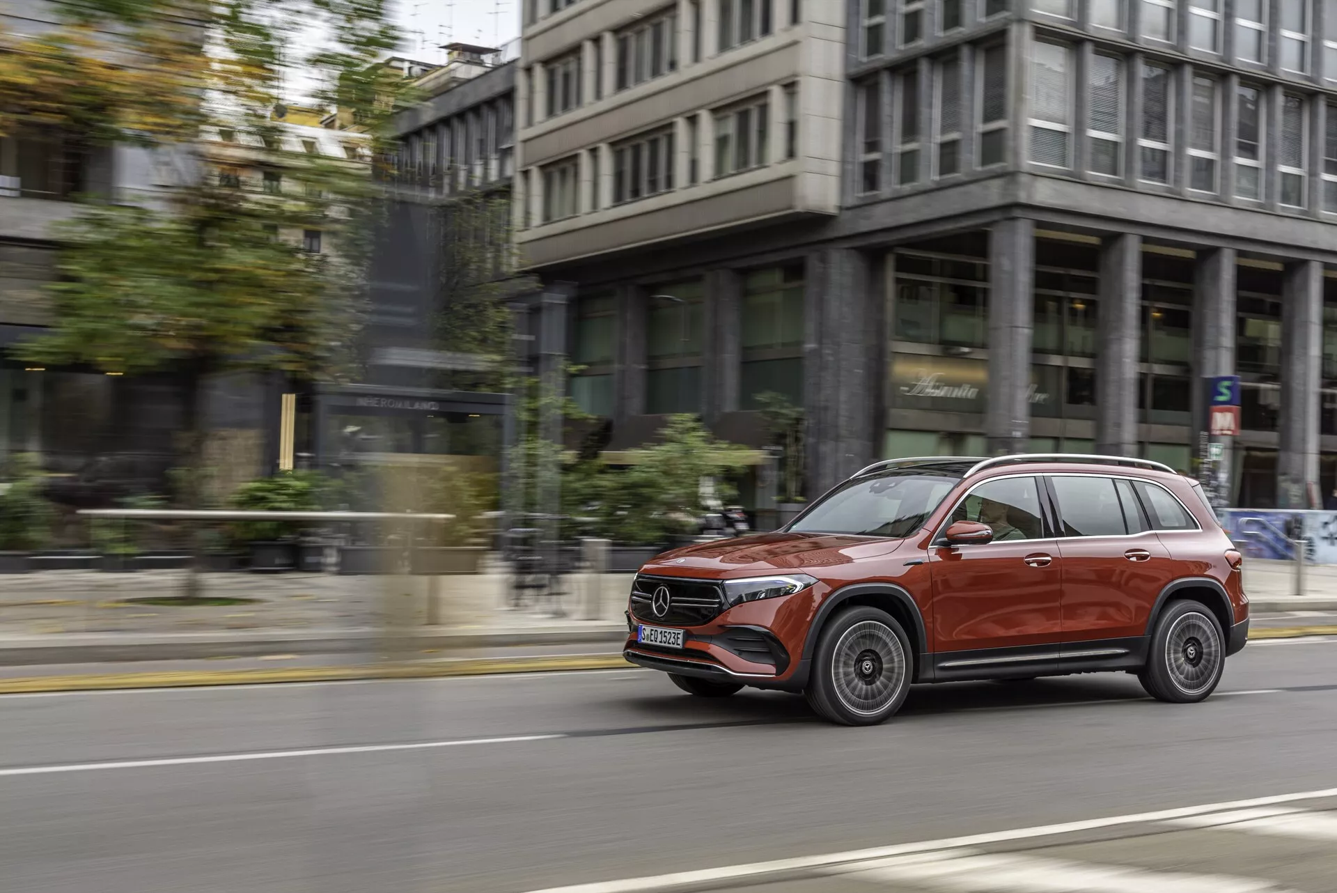 L'image montre un SUV Mercedes EQB 2021 rouge en mouvement, capturé du côté droit dans une prise de vue dynamique en panoramique. Le SUV, roulant sur une rue de la ville avec des bâtiments et un trottoir en arrière-plan, présente un extérieur brillant, des jantes en alliage élégantes, des garnitures chromées autour des fenêtres, des barres de toit et des clignotants intégrés dans les rétroviseurs latéraux. La vue légèrement inclinée vers l'avant met en évidence le côté passager avant, montrant des phares avancés sur un arrière-plan urbain flou, transmettant vitesse et élégance.