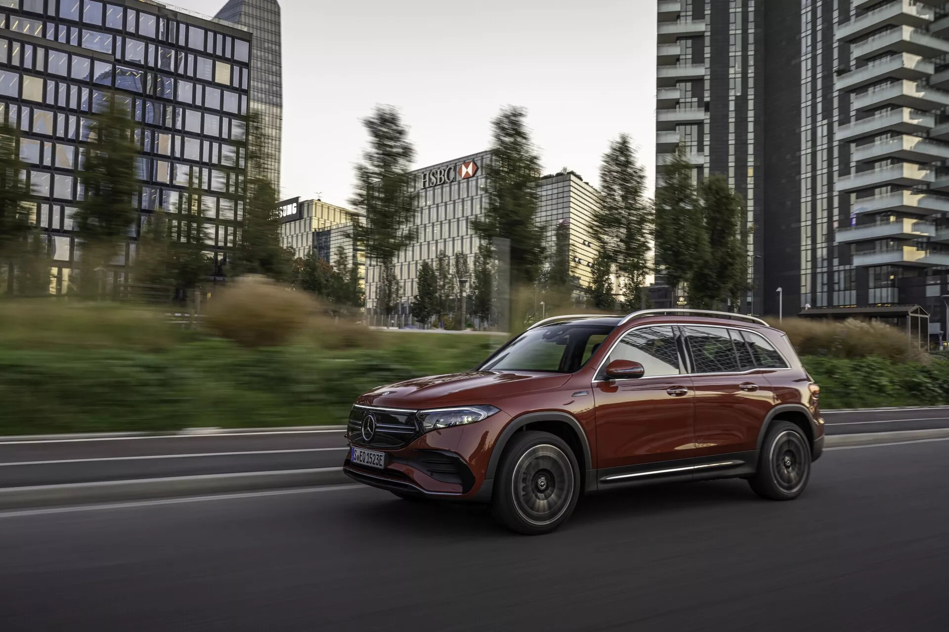 Vue dynamique latérale avant d'un SUV Mercedes EQB 2021 en mouvement sur une route urbaine avec des immeubles de bureaux en arrière-plan. Le design élégant et aérodynamique du véhicule, la finition brillante et les caractéristiques avancées telles que les phares LED modernes et les rétroviseurs latéraux avec indicateurs de direction intégrés sont mis en valeur. L'éclairage doux accentue le mélange de luxe et d'utilité de la voiture, avec un flou de mouvement sur les roues et l'arrière-plan, établissant fermement sa vitesse et sa direction de déplacement.