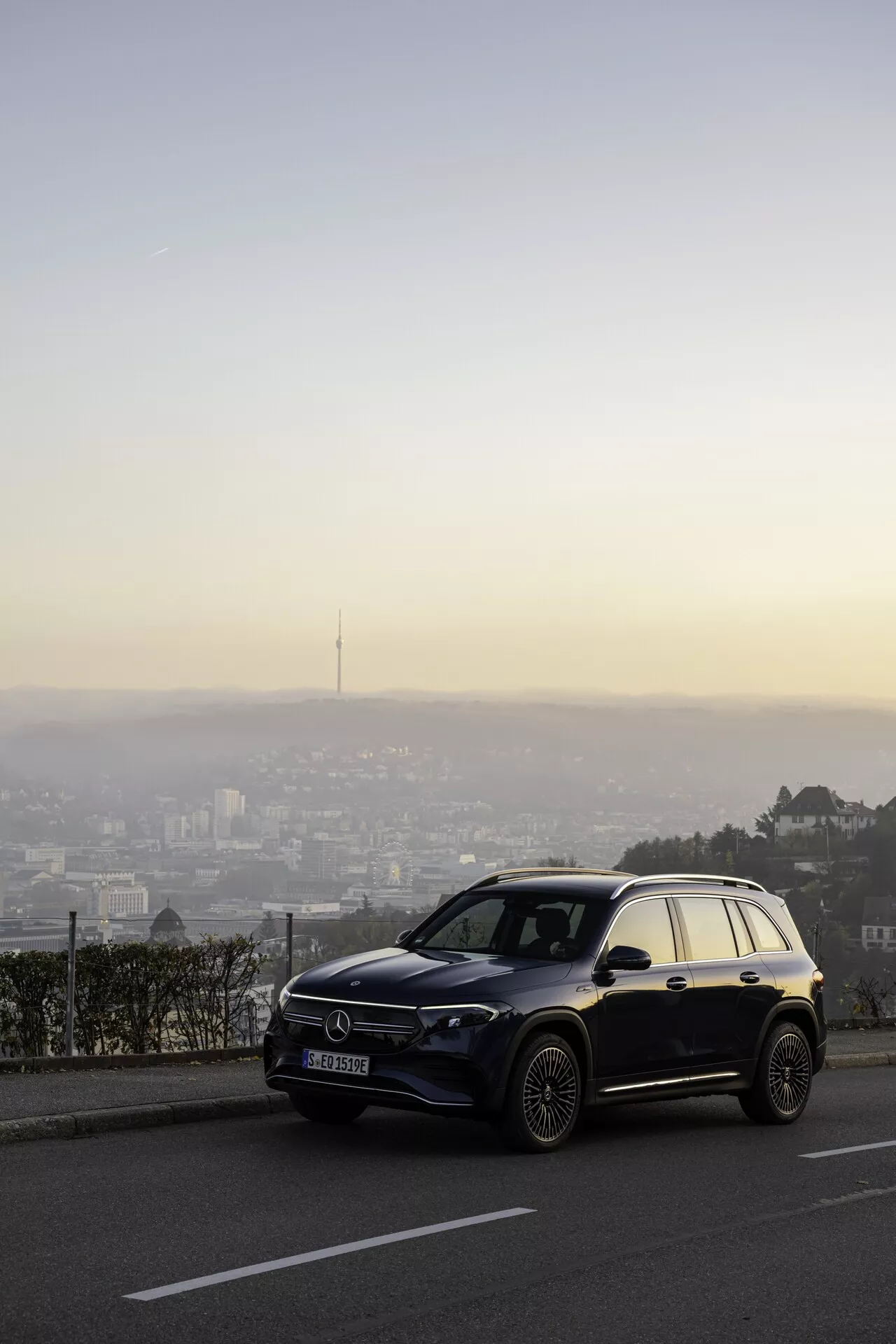Un moderne SUV Mercedes EQB 2021 positionné sur un point de vue surplombant une ville au crépuscule, capturé sous un angle de trois quarts avant montrant ses phares à LED, sa calandre avant proéminente avec un grand emblème, des jantes en alliage et une finition de peinture brillante métallique ou nacrée. Le véhicule est net contre un paysage urbain flou, placé pendant l'heure dorée avec un ciel en transition du bleu aux teintes chaudes.