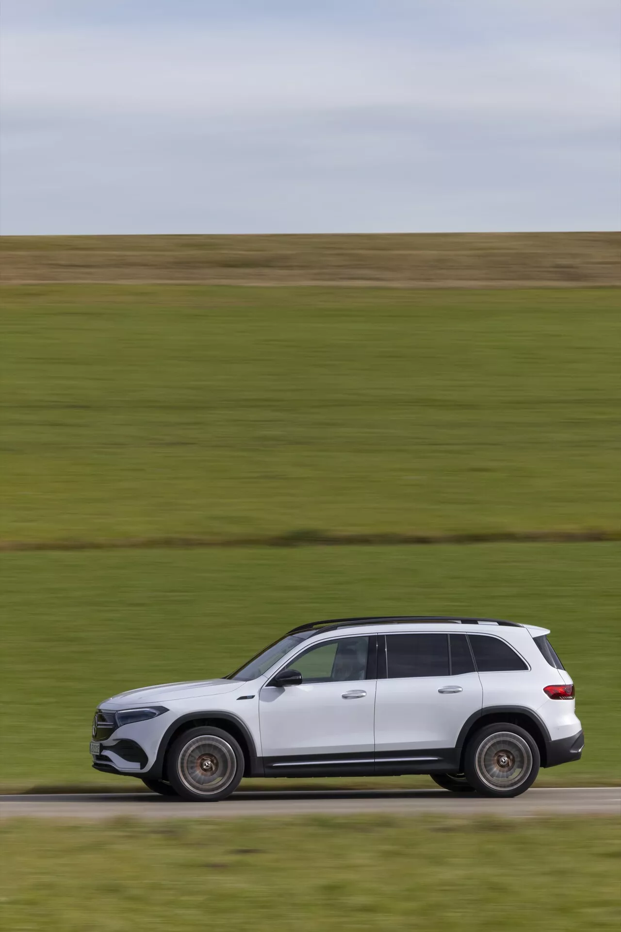 Profil latéral d'un SUV de luxe moderne en mouvement, probablement un modèle récent Mercedes EQB 2021, peint en blanc. Le véhicule, se déplaçant de gauche à droite, présente des éléments de design sophistiqués avec une carrosserie aérodynamique élégante, des roues en alliage, des garnitures chromées sur les fenêtres et des rails de toit. Malgré l'arrière-plan flou suggérant la vitesse, la voiture reste nette, mettant en valeur sa peinture impeccable et sa posture dynamique.