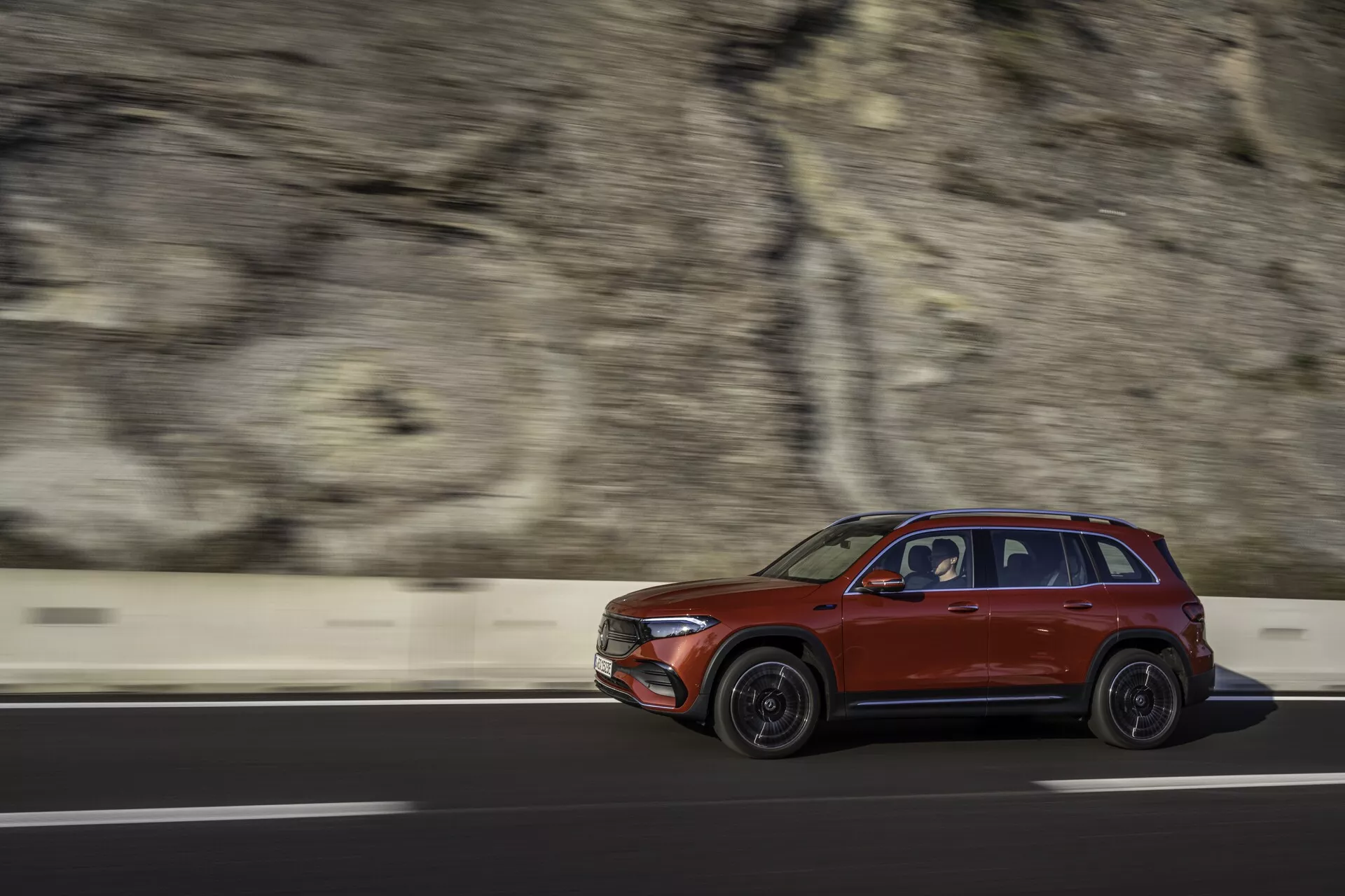 Photo dynamique de profil latéral d'un SUV Mercedes EQB 2021 rouge en mouvement, conduisant de gauche à droite sur une route avec un arrière-plan flou montrant une barrière et une paroi rocheuse. Capturée avec une vitesse d'obturation rapide, le véhicule est nettement focalisé, mettant en valeur des caractéristiques de design telles que les formes des portes, le design des roues, les rétroviseurs latéraux, les fenêtres et un porte-bagages sous une lumière douce de matin ou de fin d'après-midi.