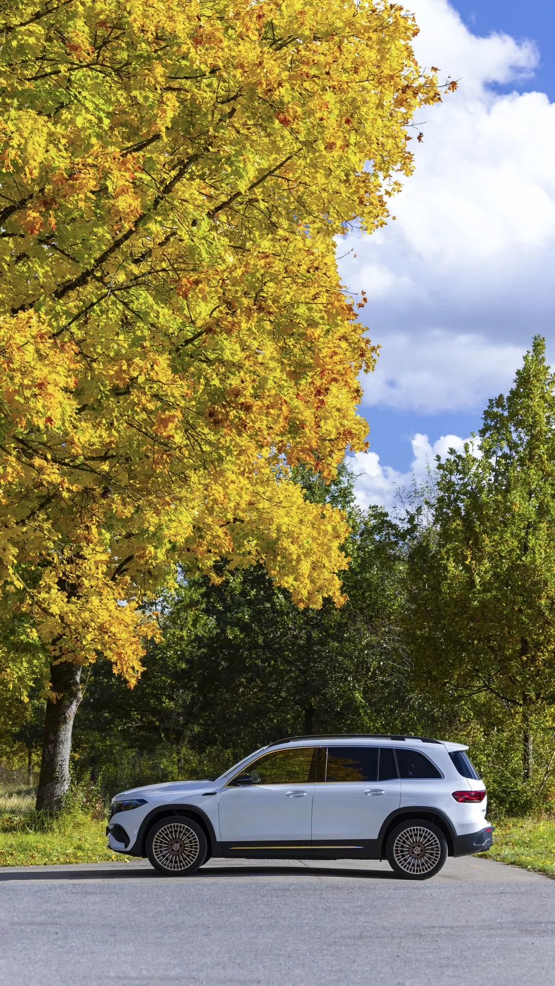 Vue de profil latéral d'un SUV de luxe Mercedes EQB 2021 garé parallèlement sur une route à deux voies, avec un schéma de couleurs bicolore avec carrosserie claire et garniture inférieure plus foncée, roues en alliage multi-rayons haut de gamme, pneus tout-terrain et entouré d'un cadre naturel serein avec des arbres au feuillage jaune, suggérant l'automne.