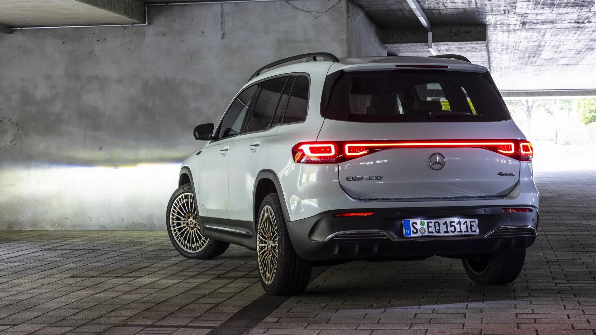 Vue arrière trois-quarts d'une Mercedes EQB 2021 blanche stationnée dans un environnement intérieur, probablement un garage. Le design arrière du véhicule, y compris le hayon, les feux arrière et le badge avec un symbole électrique, est entièrement visible. Le profil latéral du passager montre les portes arrière et une partie des portes avant, des roues optimisées aérodynamiquement avec des motifs de rayons complexes et une plaque d'immatriculation de style européen urbain. Les feux arrière illuminés mettent en valeur le design de l'éclairage. La finition blanche brillante contraste avec la structure en béton, l'éclairage artificiel et les marquages peints au sol de l'environnement intérieur.