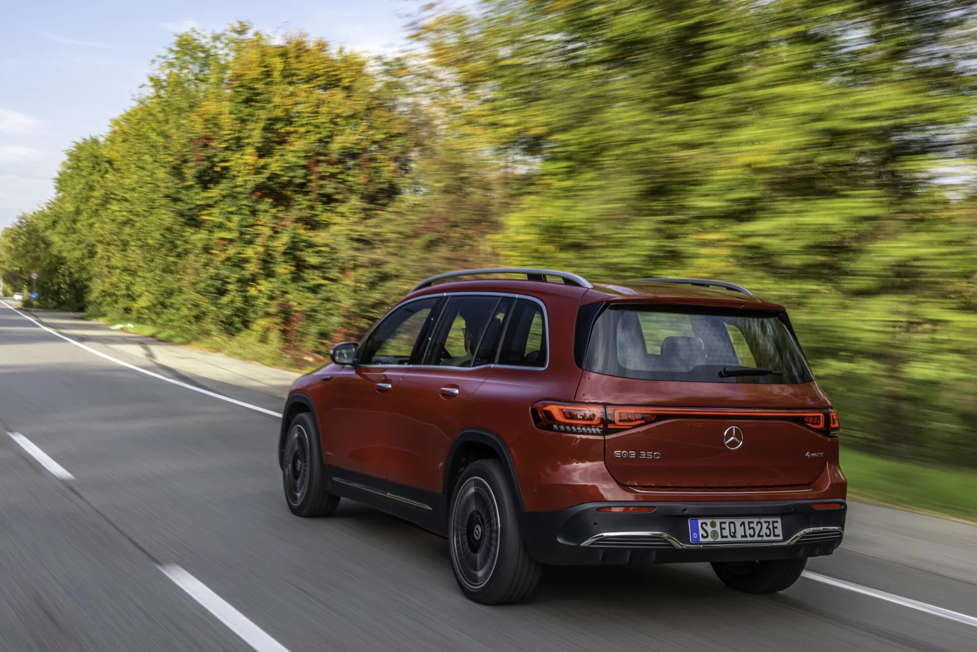 Un SUV moderne Mercedes EQB 2021 (243) en rouge métallisé capturé sous un angle de trois quarts arrière sur une route suburbaine ou rurale bien entretenue. Le design élégant et sportif de la voiture est mis en valeur par une ligne de toit inclinée, un porte-à-faux arrière court et une ligne de caractère le long du côté. Les caractéristiques visibles comprennent de grandes roues en alliage à plusieurs rayons avec des pneus à profil bas, des feux arrière à LED aérodynamiques et des capteurs de stationnement arrière. L'image véhicule une sensation de conduite dynamique avec le véhicule en mouvement et l'arrière-plan légèrement flou avec des arbres.