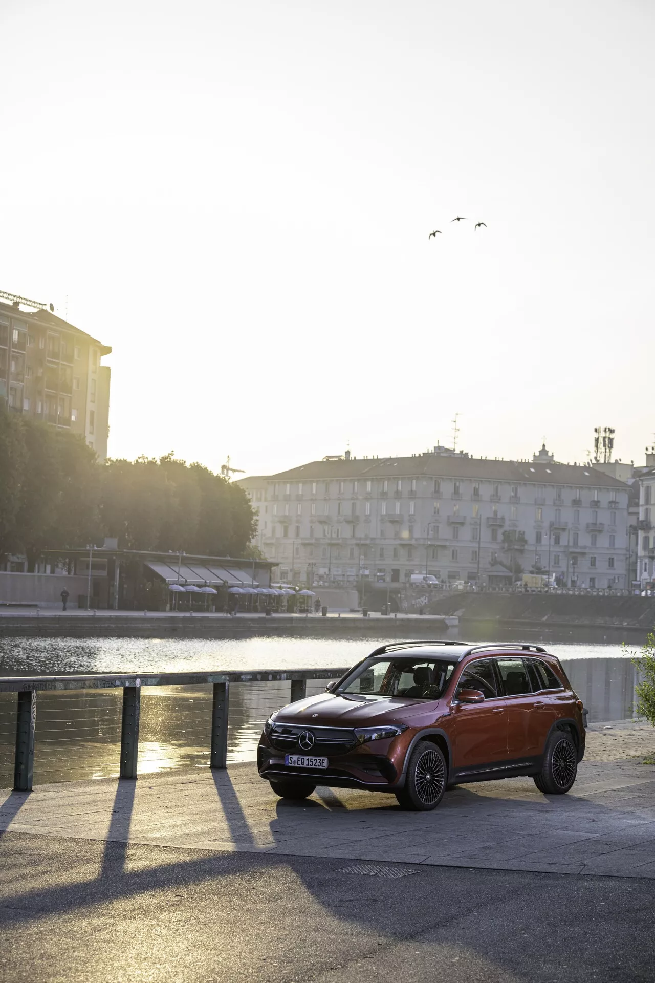 L'image présente un SUV élégant Mercedes EQB 243 de 2021 stationné au bord d'une zone riveraine urbaine. Le véhicule est capté sous un angle partiel avant et latéral, mettant en avant le côté gauche et le quart avant gauche contre un paysage aquatique serein avec des bâtiments au loin. Les éléments de design clés comme la forme des phares, le pare-chocs avant avec des feux antibrouillard intégrés ou des capteurs, le motif de la grille et les lignes du capot sont clairement visibles. La scène est baignée de lumière solaire chaude, soulignant les caractéristiques de conception supérieure du véhicule et la ligne du toit. Le pare-brise et les vitres avant permettent entrevoir l'intérieur semi-transparent de la cabine. Les systèmes d'éclairage avancés de la voiture, les capteurs dans la partie inférieure du pare-chocs avant, la hauteur de caisse élevée, les jantes en alliage et le toit panoramique suggèrent des caractéristiques de luxe et une technologie avancée. La peinture métallique reflète l'état neuf ou bien entretenu du véhicule.