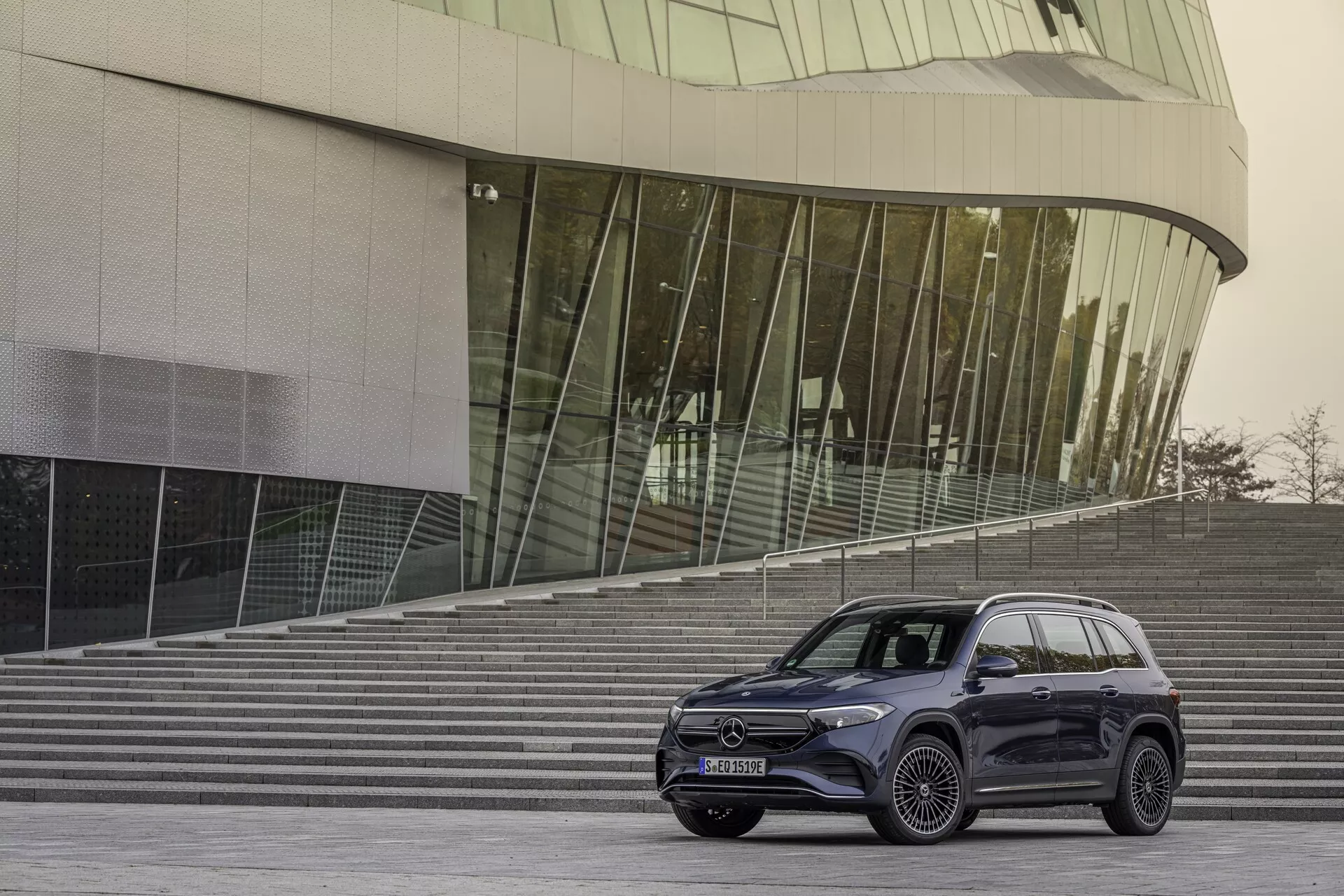 Vue frontale trois quarts d'une Mercedes EQB 2021 (243), montrant le profil latéral et la façade avant. Le véhicule, situé à 10-15 pieds de l'appareil photo, est le point central de la photographie. Il se trouve sur une zone pavée avec des marches architecturales et un bâtiment en verre moderne en arrière-plan, créant une ambiance urbaine et contemporaine. La partie avant présente des phares LED élégants, une grille proéminente avec un grand emblème et une partie du pare-chocs. Le côté montre des roues en alliage à plusieurs rayons de grand diamètre et des pneus à profil bas, indiquant une finition de luxe ou sportive. Le SUV a des lignes de carrosserie fluides et une finition sombre et brillante, probablement en graphite ou en noir. Les fenêtres sont teintées et les rétroviseurs extérieurs ont des clignotants intégrés. La ligne de toit s'incline légèrement et comporte des rails de toit pratiques. Il est probable qu'il inclut des fonctionnalités technologiques telles que des phares adaptatifs et des capteurs de stationnement, bien qu'elles ne soient pas visibles.