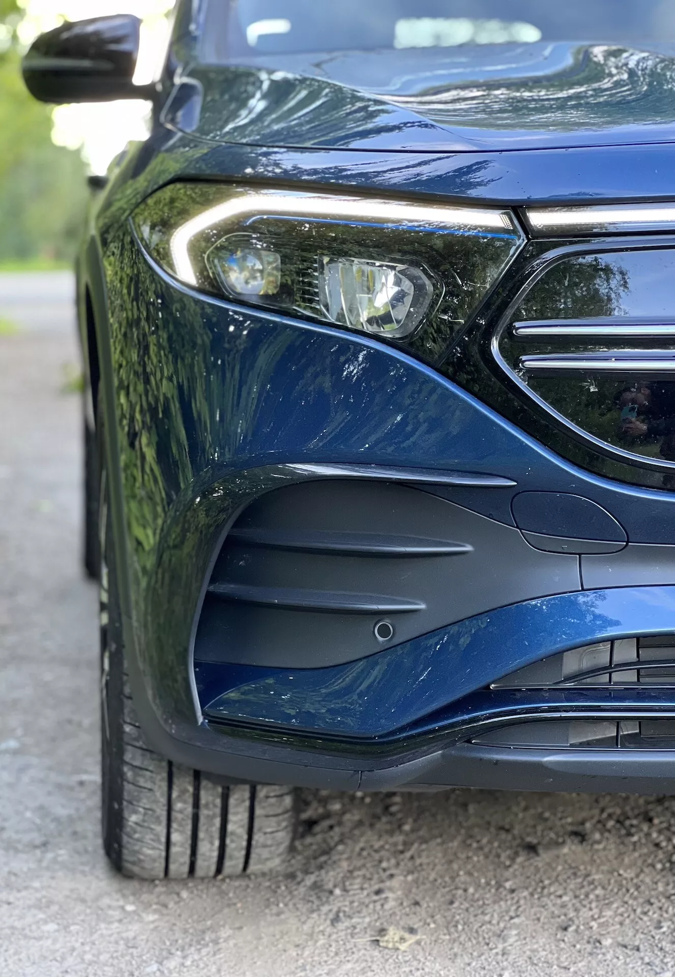 Vue rapprochée du côté avant gauche de la Mercedes-Benz EQB 2021, présentant la conception des phares LED, une partie de la calandre avec l'espace de l'emblème, le pare-chocs avant sculpté de manière aérodynamique avec des entrées d'air, une roue en alliage à multiples branches avec une bande de roulement de pneu détaillée, et une peinture métallique reflétant les courbes sophistiquées de la carrosserie