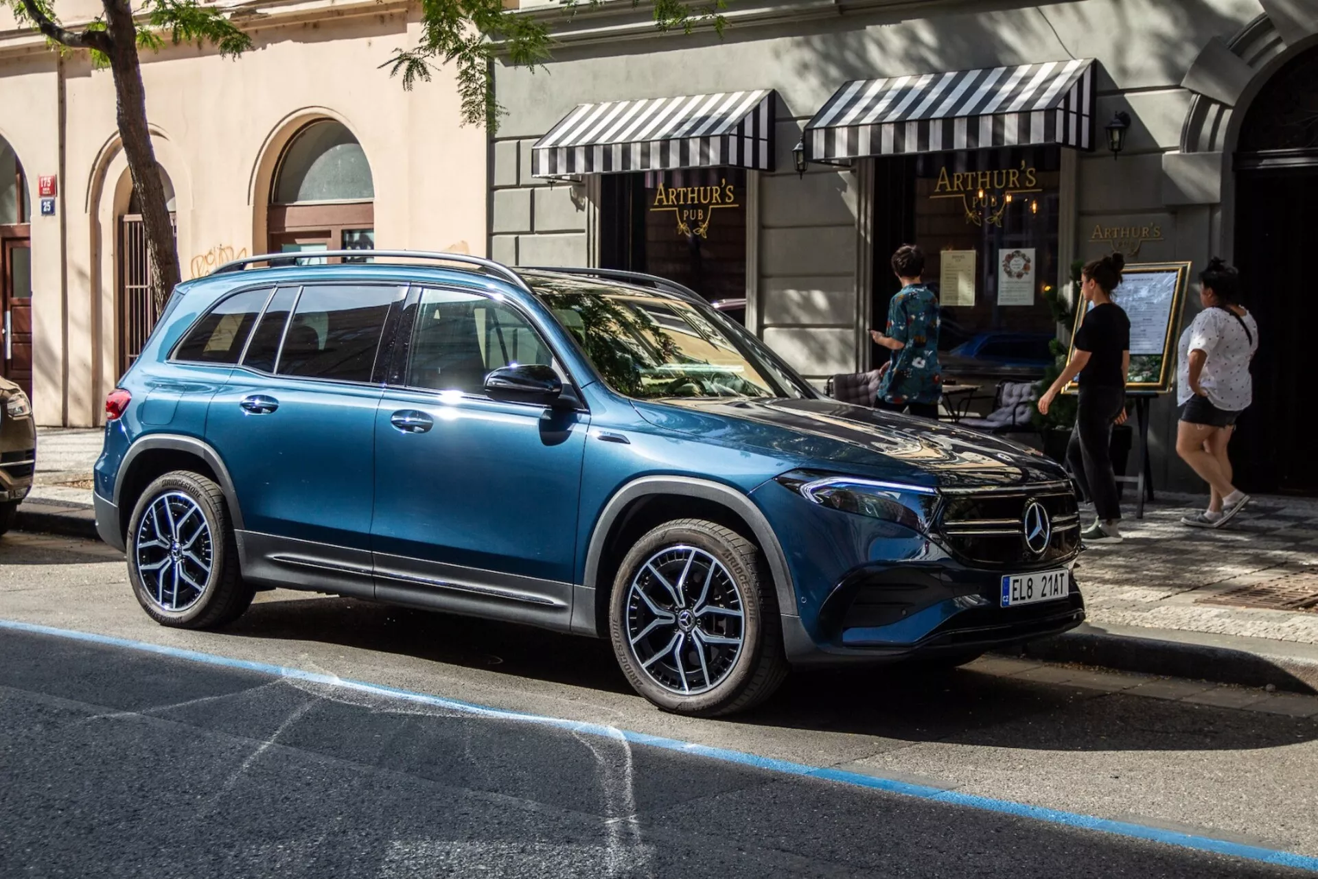 Profil latéral du Mercedes-Benz EQB 2021 dans une rue de ville, mettant en évidence le design moderne de SUV de luxe avec de grandes roues en alliage, clignotants intégrés sur les rétroviseurs latéraux, fenêtres teintées reflétant l'environnement urbain, lignes de carrosserie élégantes, rails de toit, et poignées de porte affleurantes sous un éclairage naturel