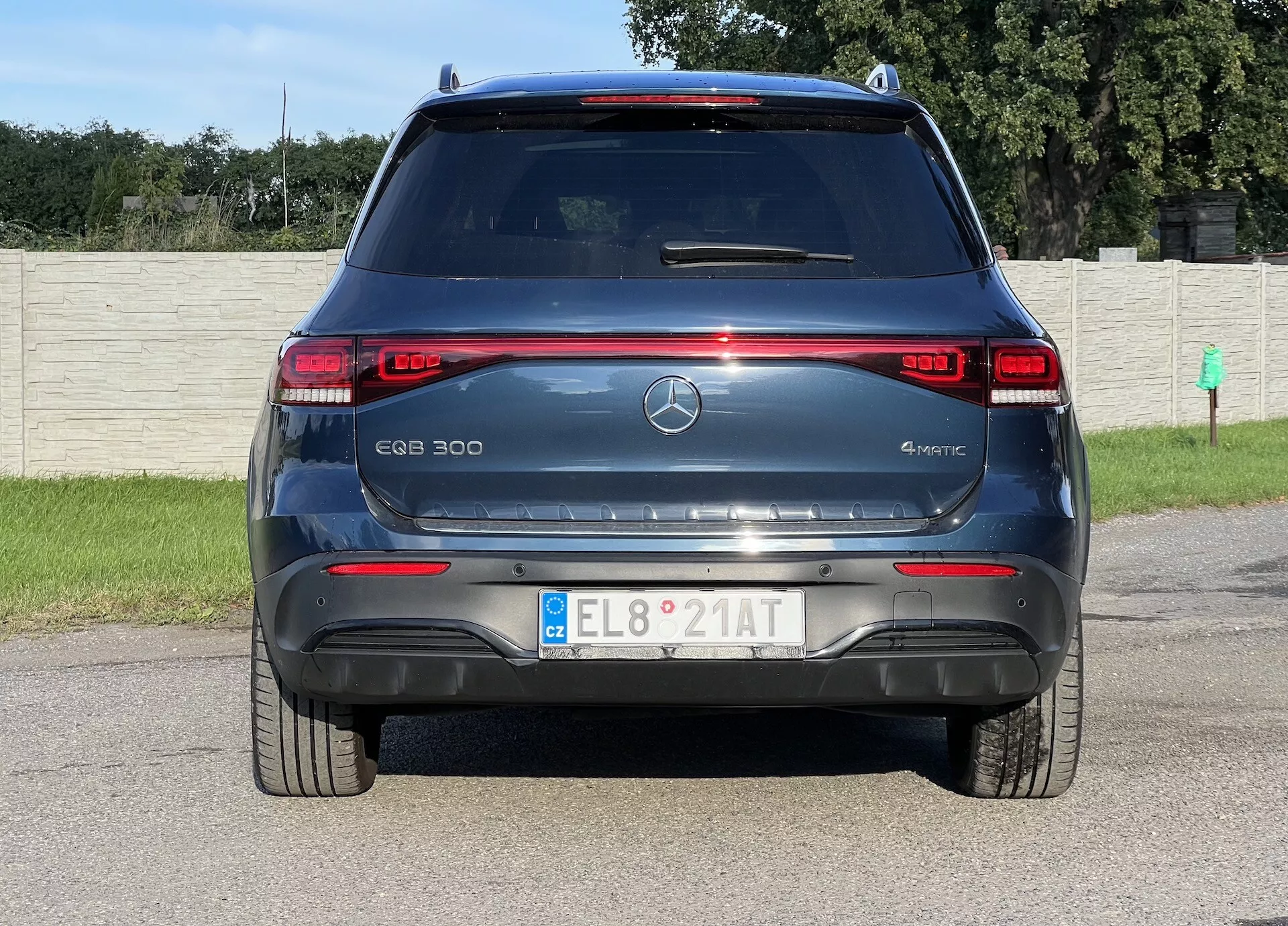 Vue arrière du SUV électrique Mercedes-Benz EQB 300 4MATIC présentant des feux arrière LED, marquage du modèle et électrique, emblème étoile à trois branches, pare-brise arrière avec dégivreur et essuie-glace, capteurs de stationnement, réflecteurs rouges, plaque d'immatriculation européenne, spoiler arrière aérodynamique et système de traction intégrale 4MATIC, sur fond de champ d'herbe et clôture.