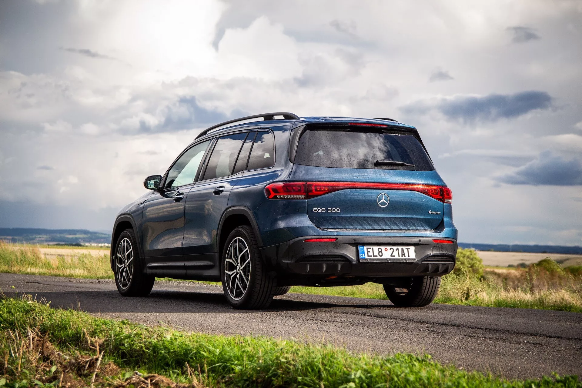 SUV électrique Mercedes-Benz EQB 2021 présenté sous un angle arrière dynamique de trois quarts mettant en évidence le design élégant du véhicule, y compris les feux arrière combinés, un spoiler de toit sportif, des jantes en alliage et le badge EQB sur fond de ciel couvert et de paysage rural.