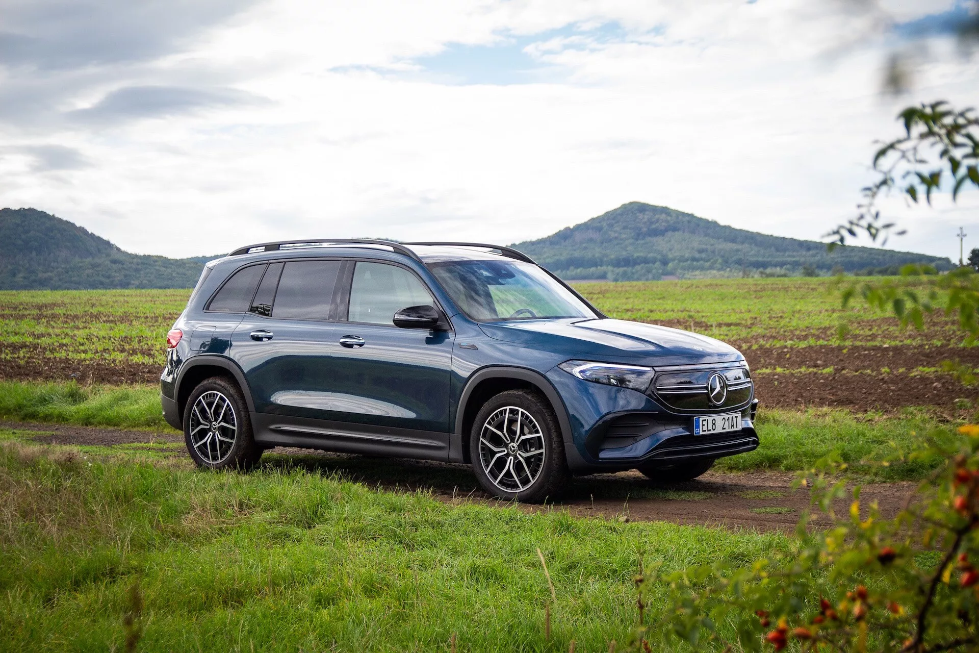 Profil latéral d'un SUV Mercedes-Benz EQB 2021 de couleur foncée garé sur un terrain herbeux avec une piste de terre visible à l'avant, mettant en valeur des jantes en alliage, des barres de toit et des lignes de design sportives sur fond de paysage rural avec un ciel partiellement nuageux
