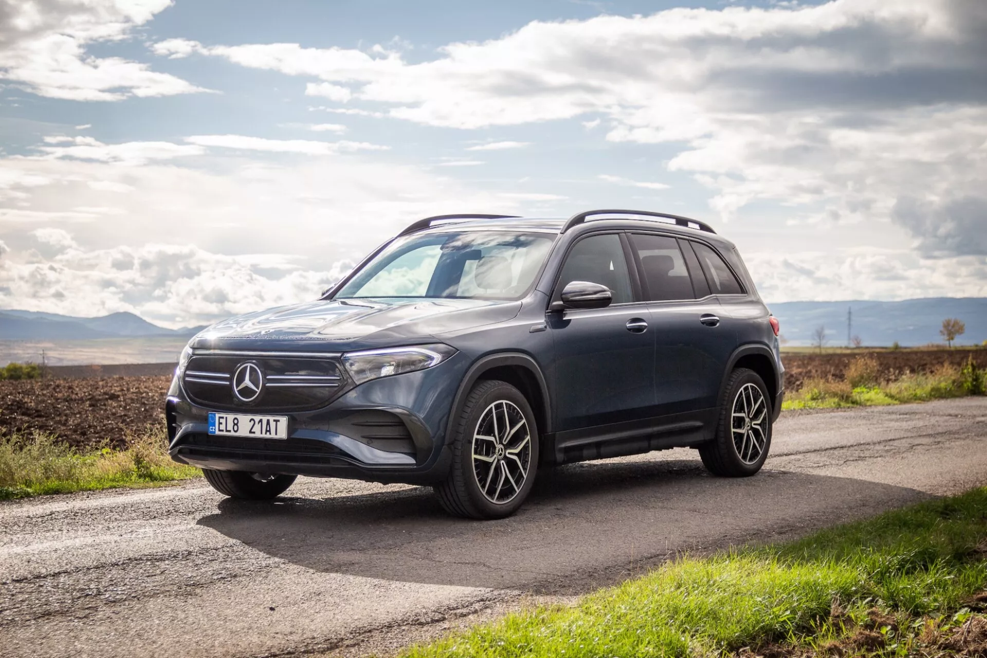 Vue latérale et frontale du SUV de luxe Mercedes-Benz EQB 2021 présentant un design aérodynamique, des phares LED, des roues en alliage à multiples rayons et une garde au sol élevée sur une route d'asphalte sous un ciel partiellement nuageux
