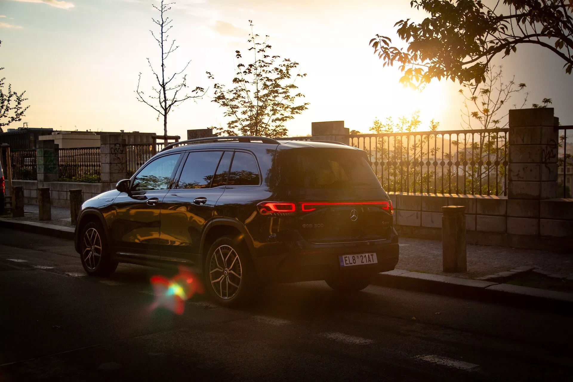 Mercedes-Benz EQB 2021 dans une vue arrière dynamique des trois quarts au coucher du soleil avec éblouissement de l'objectif, mettant en avant son design moderne de SUV, feux arrière LED, garé dans une rue de la ville reflétant la lumière chaude du soleil sur sa peinture sombre et brillante et jantes en alliage à plusieurs branches, y compris les capteurs de stationnement visibles sur le pare-chocs arrière