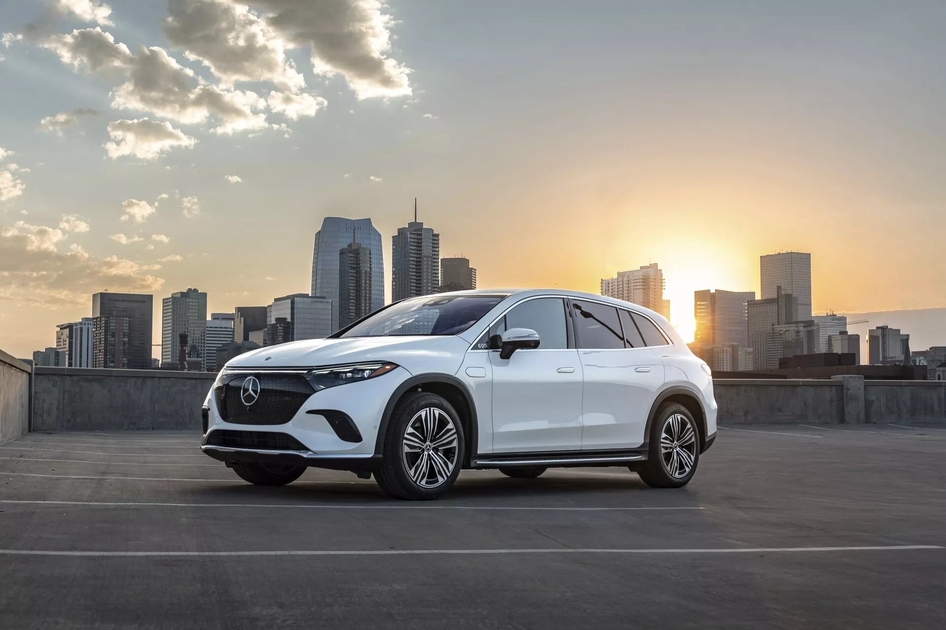 Un sophistiqué Mercedes EQS-SUV 2022 blanc garé avec une ligne d'horizon de ville et un soleil couchant en arrière-plan. L'angle de vue de trois quarts avant met en valeur la grille distinctive et proéminente du véhicule avec un motif en maille détaillé et un emblème central, des phares LED élégants, des doubles prises d'air ou ventilations et de grandes roues en alliage multi-branches. La ligne de toit doucement inclinée et l'augmentation de la hauteur de caisse indiquent son design de SUV. Les rétroviseurs latéraux abritent probablement des fonctionnalités technologiques telles que des clignotants intégrés ou des caméras pour la surveillance des angles morts. L'image est magnifiquement illuminée avec des reflets doux du soleil couchant, accentuant les lignes de conception de la voiture. La toile de fond urbaine avec des gratte-ciel ajoute une touche contemporaine et sophistiquée, laissant entendre un public cible de consommateurs urbains qui apprécient le style et la technologie avancée.