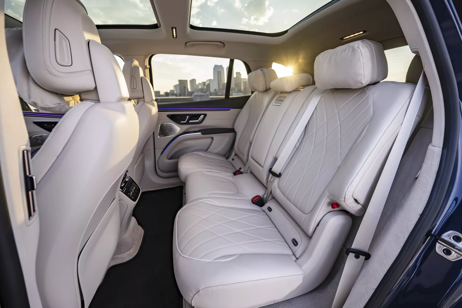 Vue intérieure du Mercedes EQS-SUV 2022 montrant le compartiment passager arrière luxueux. L'image, prise sous un angle légèrement incliné vers le côté droit de la voiture, révèle des sièges en cuir de haute qualité blanc ou gris clair avec un motif à losanges piqué. Avec trois appuie-têtes, les sièges offrent un espace généreux, indiquant une attention particulière au confort et à l'élégance. On peut également voir la partie arrière du siège passager avant, également habillée de cuir haut de gamme. L'éclairage d'ambiance sophistiqué avec une douce lueur violette le long des panneaux de porte et de l'accoudoir central renforce la sensation premium. L'accoudoir central est équipé de commandes, probablement pour les réglages climatiques et de siège, et comprend deux porte-gobelets.