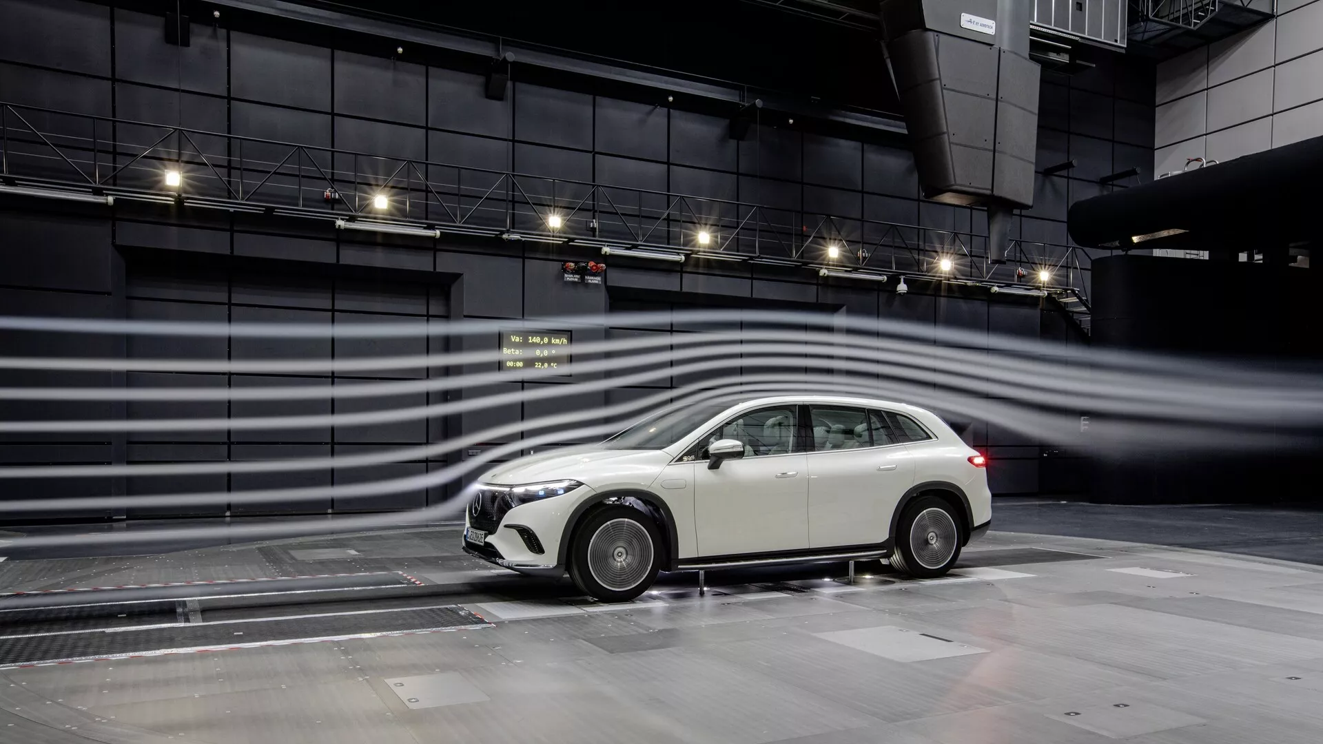 L'image présente un profil latéral du Mercedes EQS-SUV 2022, mettant en évidence son design moderne dans une installation de test contrôlée. Le modèle hatchback ou break du véhicule est accentué par une calandre avant proéminente, des rétroviseurs latéraux élégants, des jantes alliage multibranches, des phares antibrouillard intégrés dans le pare-chocs avant et une antenne montée sur le toit. Les lignes aérodynamiques et les stries visualisant le flux d'air autour de la voiture suggèrent un accent mis sur l'efficacité énergétique et les performances, avec la surface mouillée reflétant la voiture, possiblement indiquant des tests d'interaction avec la surface de la route. La configuration suggère des technologies avancées et des tests aérodynamiques, potentiellement pour des fonctionnalités telles que l'aérodynamique active, les systèmes d'assistance à la conduite et le contrôle de traction.