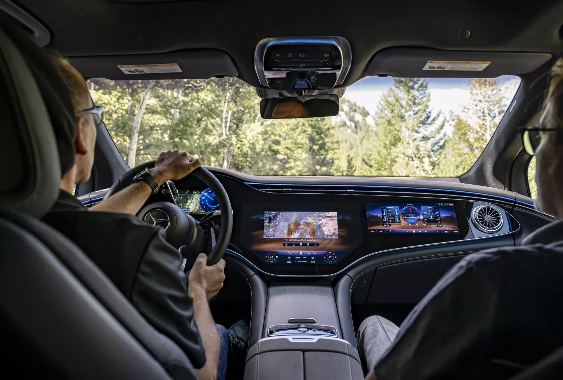 Vue intérieure d'une Mercedes EQS-SUV 2022 depuis la perspective du siège central arrière montrant un tableau de bord moderne avec écran numérique continu, volant contemporain avec commandes intégrées, sièges en cuir de couleur claire luxueux et une console centrale surélevée. Le pare-brise offre une vue d'une route entourée de forêts, suggérant que la voiture est en mouvement.