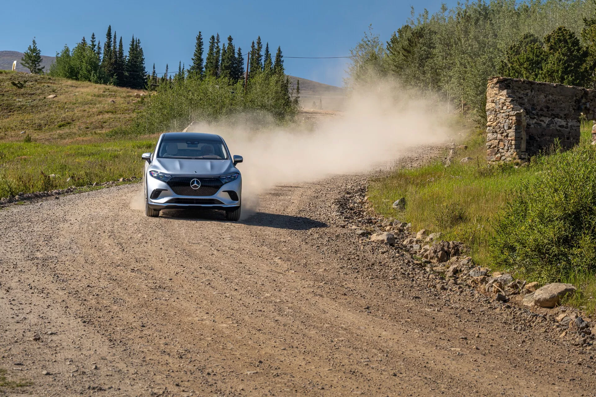 Mercedes EQS SUV 2022 – Un hatchback moderne ou SUV compact roulant vers la caméra sur une route en terre, créant une traînée de poussière. La voiture est positionnée au centre, affichant de manière proéminente une calandre avant, des phares, un capot, un pare-brise et une partie du toit. L'environnement suggère un cadre rural ou hors route avec de l'herbe de chaque côté, un ciel clair et les vestiges d'une ancienne structure en pierre en arrière-plan.