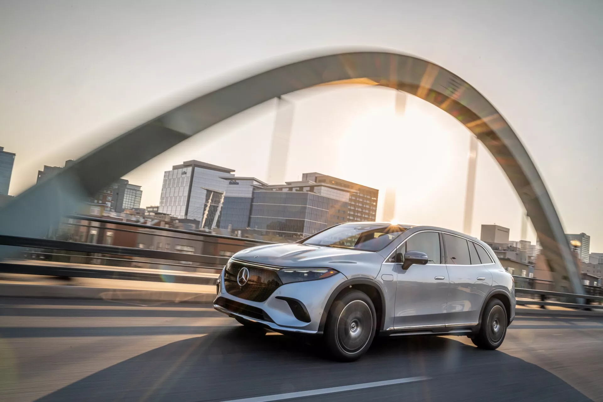 L'image montre un Mercedes EQS-SUV 2022 argenté roulant sur un pont surélevé. Capturée sous un angle bas, la vue de trois quarts avant met en valeur le design moderne du SUV. Le côté conducteur révèle une calandre chromée proéminente avec un emblème, des lignes élégantes, des phares LED stylisés et un léger flou de mouvement sur les roues indiquant la vitesse. Le flare du soleil et la silhouette floue d'une ville ajoutent un effet dramatique, soulignant le luxe et l'ingénierie avancée.
