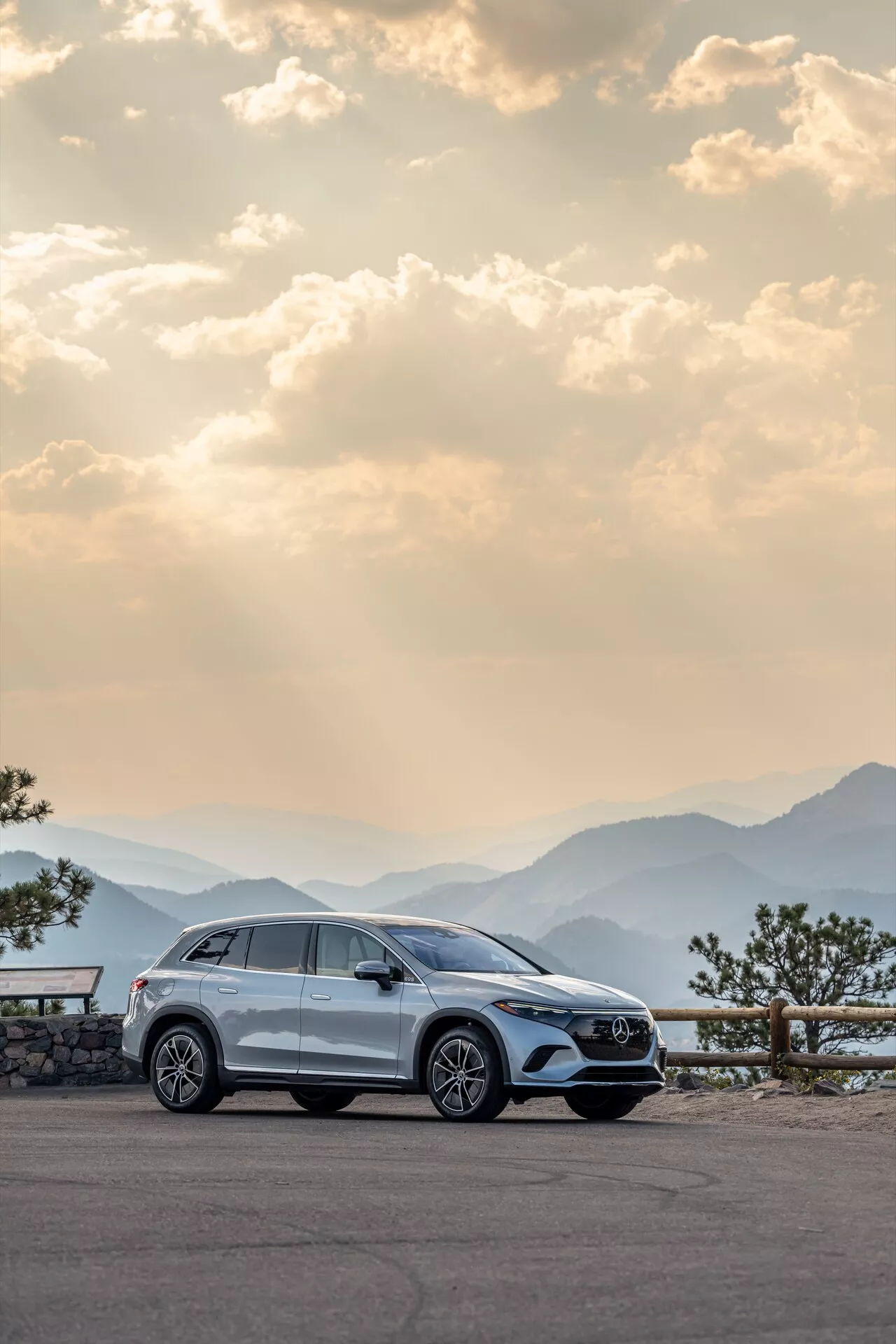 Le SUV moderne Mercedes EQS-SUV 2022 est garé sur un point de vue pittoresque, mettant en valeur son profil latéral droit avec des jantes en alliage, des poignées de porte, des rétroviseurs latéraux, des garnitures de fenêtre et des roues légèrement tournées vers la gauche. Capturé à l'aube ou au crépuscule avec une lumière chaude et diffuse, en arrière-plan des chaînes de montagnes panoramiques et d'un ciel légèrement nuageux, mettant en avant le luxe, l'aventure et la haute qualité artisanale.