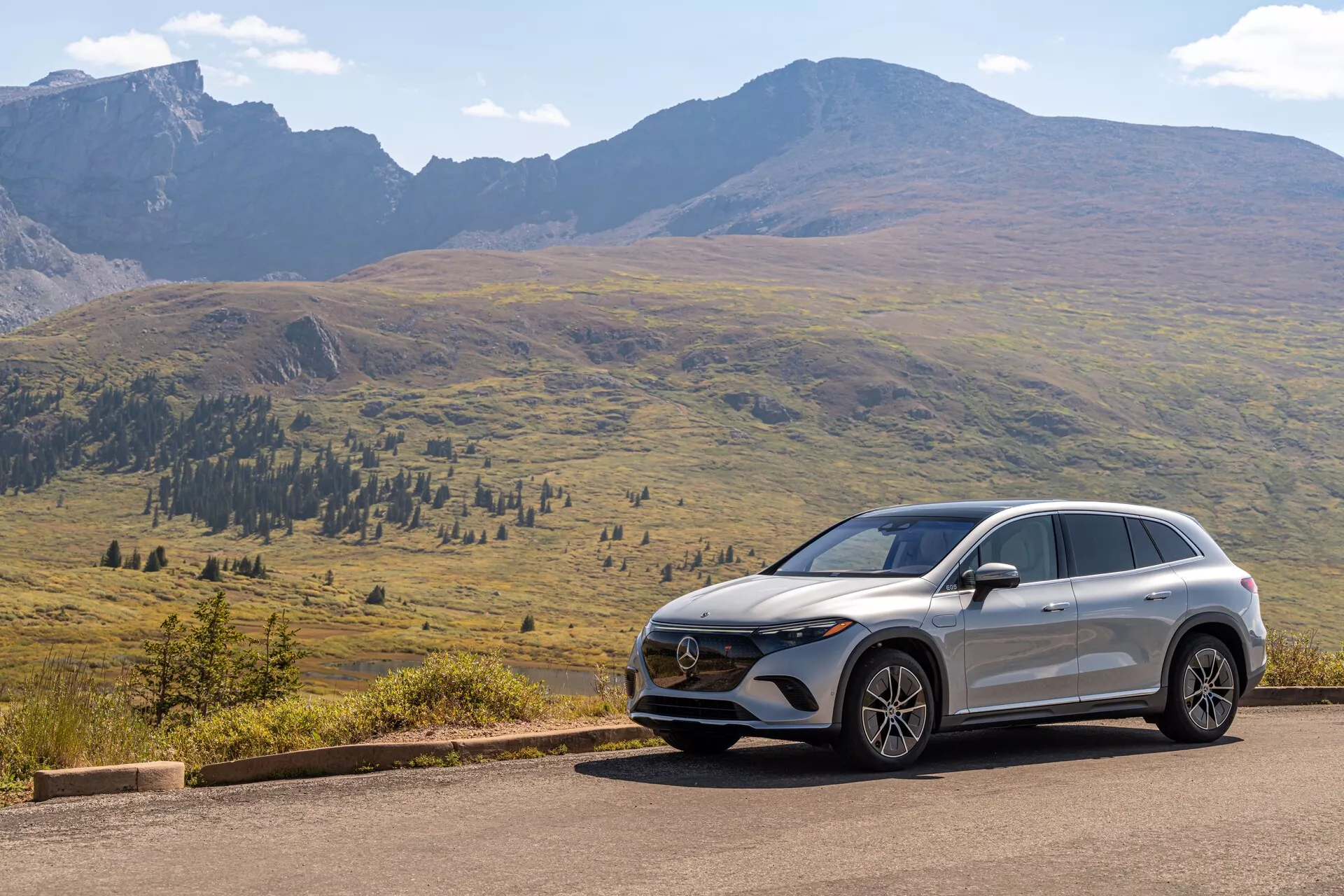 L'image montre un Mercedes EQS-SUV 2022 moderne positionné sur une route asphaltée avec un paysage montagneux pittoresque en arrière-plan sous un ciel dégagé avec quelques nuages dispersés. La vue de trois quarts avant révèle des éléments clés tels que la calandre ornée de l'emblème, l'assemblage avancé des phares et la conception du pare-chocs avant avec des capteurs ou des caméras. Les lignes aérodynamiques sur le capot et les côtés, ainsi que le profil latéral montrant les jantes en alliage, les rétroviseurs, les lignes de caractère et la pente du toit, mettent en avant les caractéristiques de design et de technologie du véhicule.
