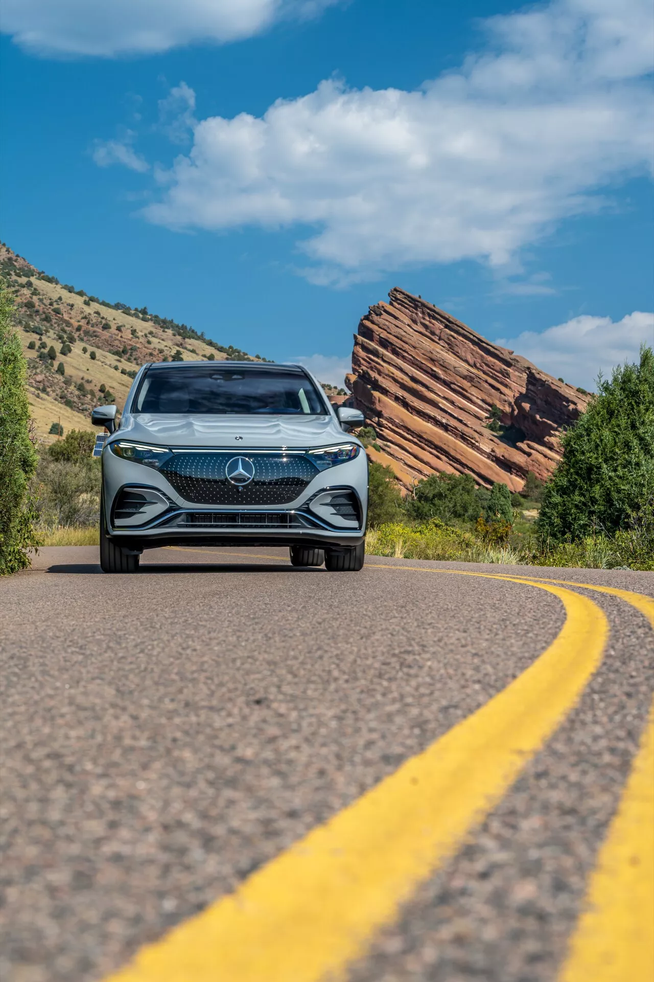 Prise en angle bas d'un Mercedes EQS SUV 2022 s'approchant d'une route sinueuse, présentant la calandre audacieuse avec un grand emblème étoile, un design en diamant complexe, des phares LED et des feux de jour, sur fond de ciel bleu clair et de terrain naturel accidenté avec des formations rocheuses stratifiées.