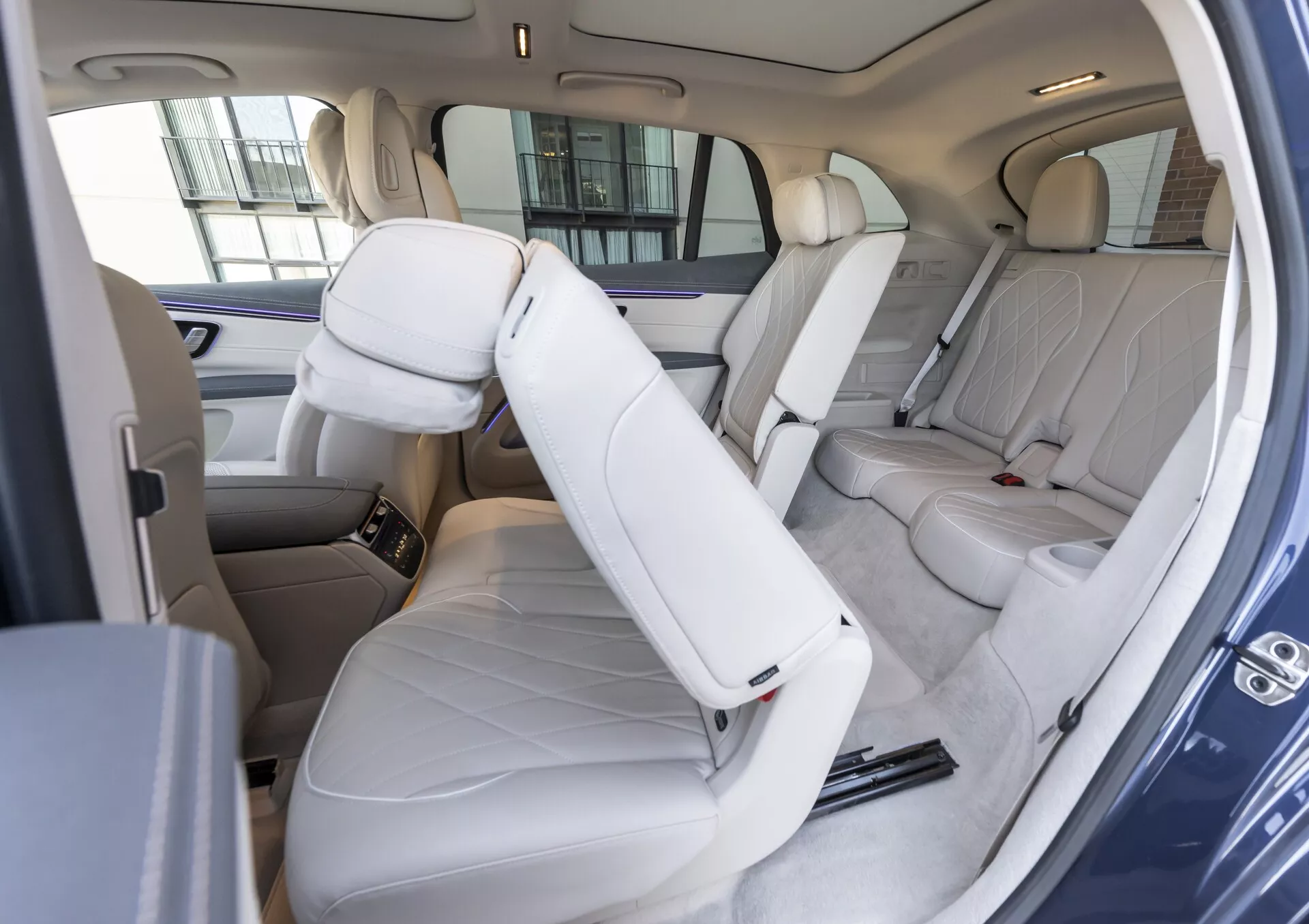L'image capture l'intérieur d'un Mercedes EQS SUV 2022, en se concentrant sur le compartiment passager arrière vue de côté avec un léger angle vers le bas. Les sièges en cuir léger à l'arrière et en partie à l'avant sont visibles, ce qui indique le luxe. L'accoudoir central à l'arrière est relevé, avec un aperçu de la console centrale avancée entre les sièges avant. Les caractéristiques de sécurité comme les ceintures de sécurité et les appuie-tête sont évidentes. L'habitacle spacieux avec un espace généreux pour les jambes et la tête souligne le confort, et le panneau de porte intègre des commandes pour les vitres électriques ou les serrures.