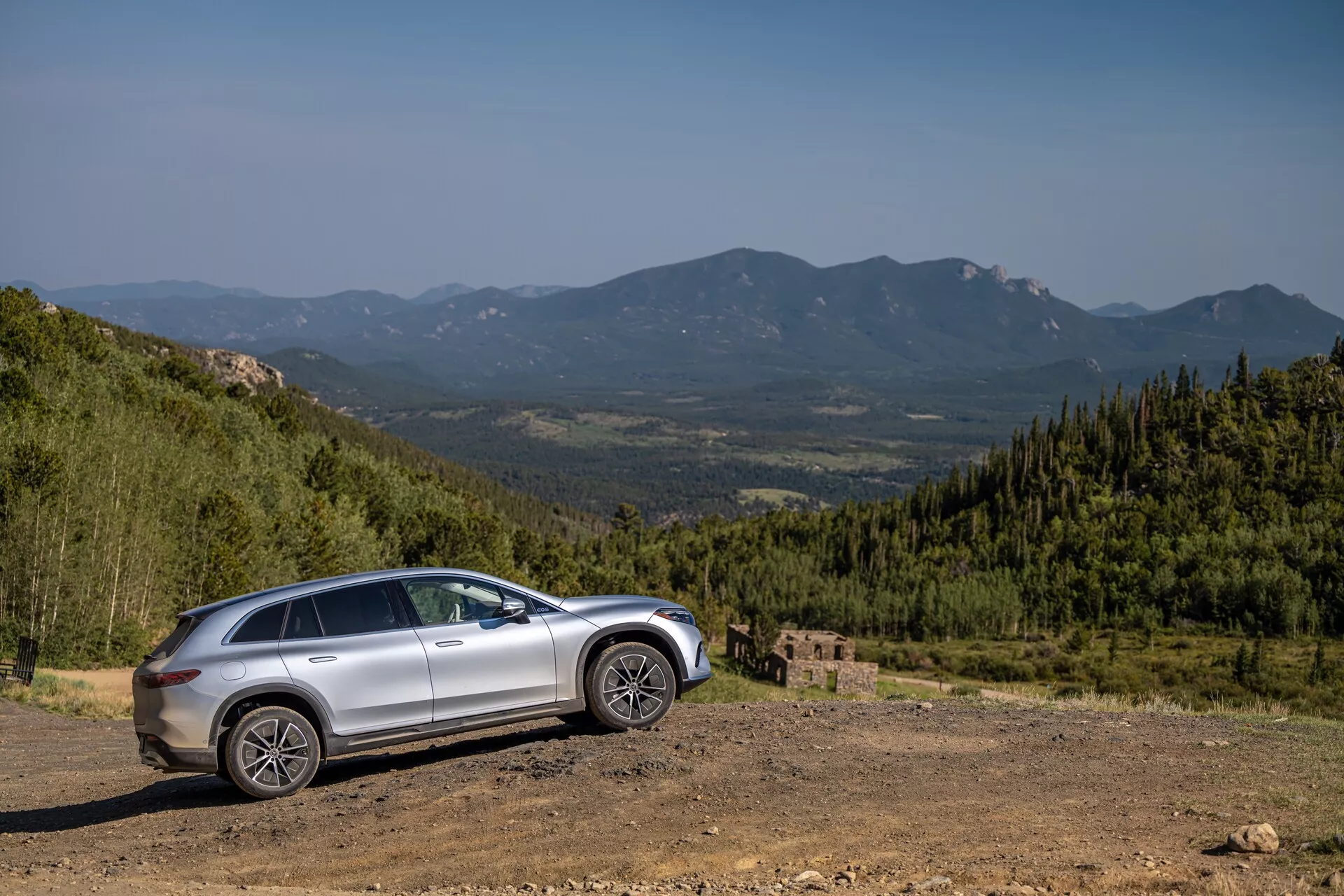 Profil latéral d'un Mercedes EQS-SUV argenté de 2022 sur un terrain accidenté avec une toile de fond montagneuse pittoresque, capturé sous un angle de 30 à 40 degrés. Les caractéristiques incluent une garde au sol élevée, une hauteur de conduite surélevée, de grandes roues robustes, des lignes sculptées sur les portes, un possible toit ouvrant, des rétroviseurs latéraux avec clignotants intégrés, des fenêtres teintées et des phares et feux arrière à LED modernes.