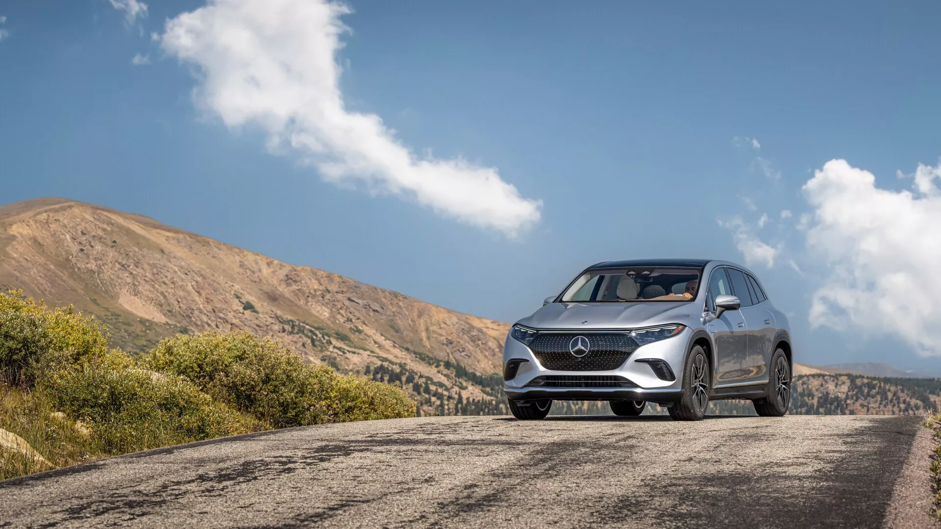 SUV argenté garé sur une route de montagne avec vue panoramique en arrière-plan, représenté en vue avant trois-quarts basse. La calandre avant, l'emblème et les phares LED modernes de la voiture sont mis en avant, ainsi que des lignes de design détaillées sur la partie inférieure avant. La peinture argentée reflète la lumière ambiante, mettant en valeur les courbes et les éléments aérodynamiques. Des jantes premium et des pneus toutes saisons sont visibles, suggérant un niveau de finition supérieur. Le contexte général implique des capacités adaptées à un mode de vie actif.