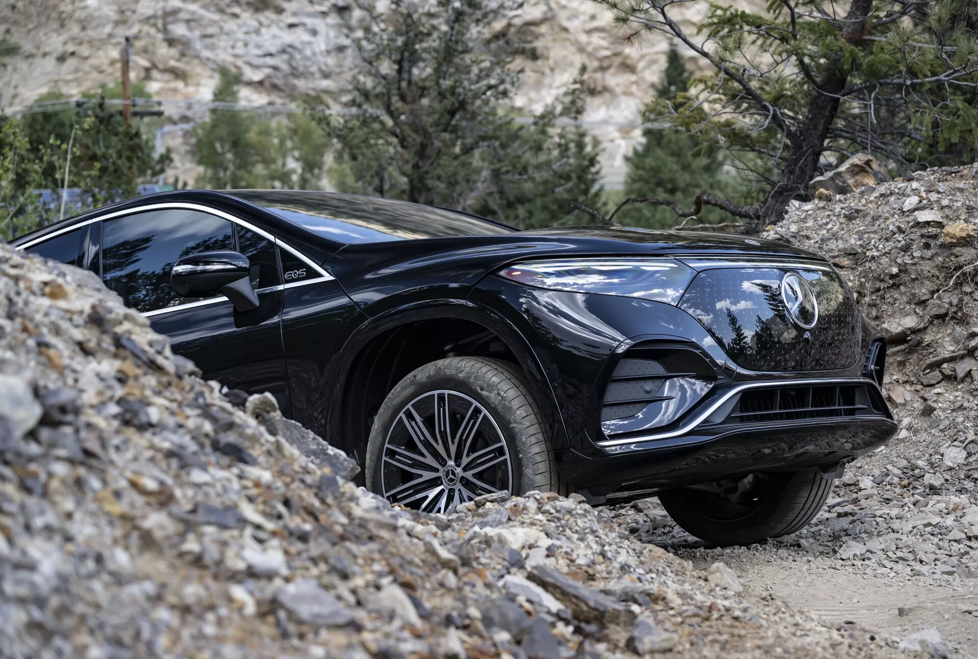 Image d'un Mercedes EQS-SUV 2022 noir négociant un terrain tout-terrain vu de profil bas. L'accent est mis sur le quart avant du véhicule, montrant des détails tels que la finition noire brillante, les lignes élégantes, la calandre avant avec emblème, les phares distinctifs, les arches de roue musclées, les jantes en alliage et la bande de roulement adéquate pour tous les terrains. La roue avant est légèrement tournée vers la droite, indiquant une manœuvre. L'arrière-plan naturel avec des rochers et une végétation clairsemée met en évidence la capacité tout-terrain du SUV.