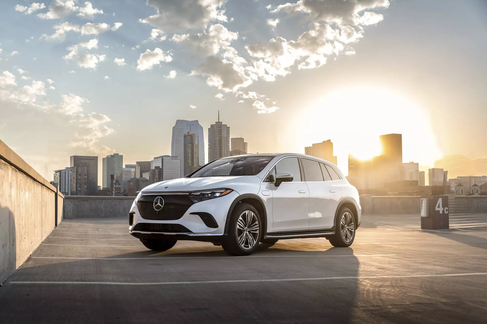 L'image montre un SUV moderne Mercedes EQS-SUV 2022 positionné dans un parking en plein air avec la ligne d'horizon de la ville en arrière-plan, capturé au lever ou au coucher du soleil avec un éclairage arrière chaleureux. La vue frontale trois quarts met en évidence la calandre, les phares, le design du pare-chocs avant, le capot et le côté du conducteur, prise sous un angle bas près du niveau des roues. Les détails incluent une calandre proéminente avec un grand emblème, des phares LED élégants, une caméra frontale ou un groupe de capteurs, des jantes en alliage multi-branches, des clignotants intégrés aux rétroviseurs latéraux, des lignes de contour subtiles pour l'aérodynamisme et une ligne de toit légèrement inclinée. La composition de la photo positionne la voiture à droite du cadre avec la ligne d'horizon de la ville et le soleil à gauche, ajoutant un effet de lens flare et un attrait artistique, adaptée pour des contextes automobiles, de luxe et de style de vie urbain.