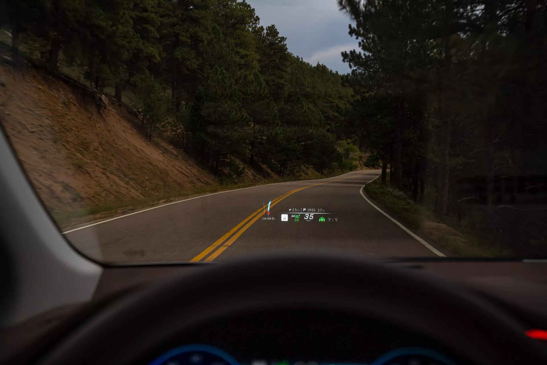 Vue intérieure d'une Mercedes EQS-SUV 2022 depuis le siège du conducteur, montrant une route à deux voies non divisée qui tourne à gauche, encadrée par des arbres et une colline. Le tableau de bord est visible avec un segment du volant éclairé en bleu, un cluster d'instruments digital et un affichage tête haute (HUD) projetant la vitesse (35 mph) et les instructions de navigation sur le pare-brise.