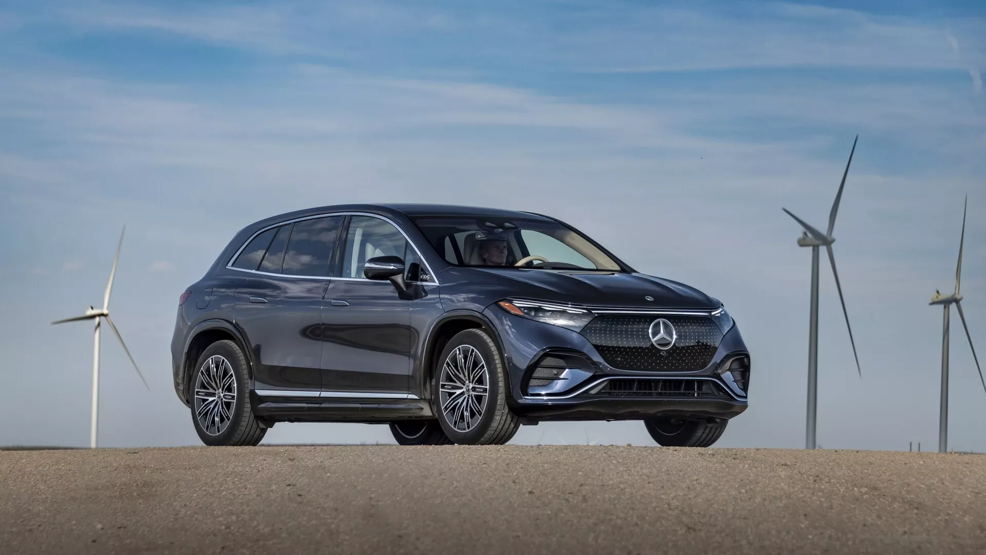 Profil latéral du Mercedes EQS SUV 2022 de couleur foncée, garé sur une surface plane. L'image, prise sous un léger angle avant, montre un design élégant et moderne avec des lignes courbes et des surfaces lisses mettant en valeur l'efficacité aérodynamique. Les caractéristiques notables incluent une calandre proéminente avec un logo de marque central, des phares LED élégants, des prises d'air ou des capteurs dans le pare-chocs inférieur, des roues en alliage à multiples rayons sophistiquées, des rétroviseurs latéraux avec clignotants intégrés, des fenêtres encadrées par du chrome et une ligne de toit qui descend doucement avec des barres de toit. A l'arrière-plan, des éoliennes, suggérant la nature écologique ou électrifiée du véhicule.