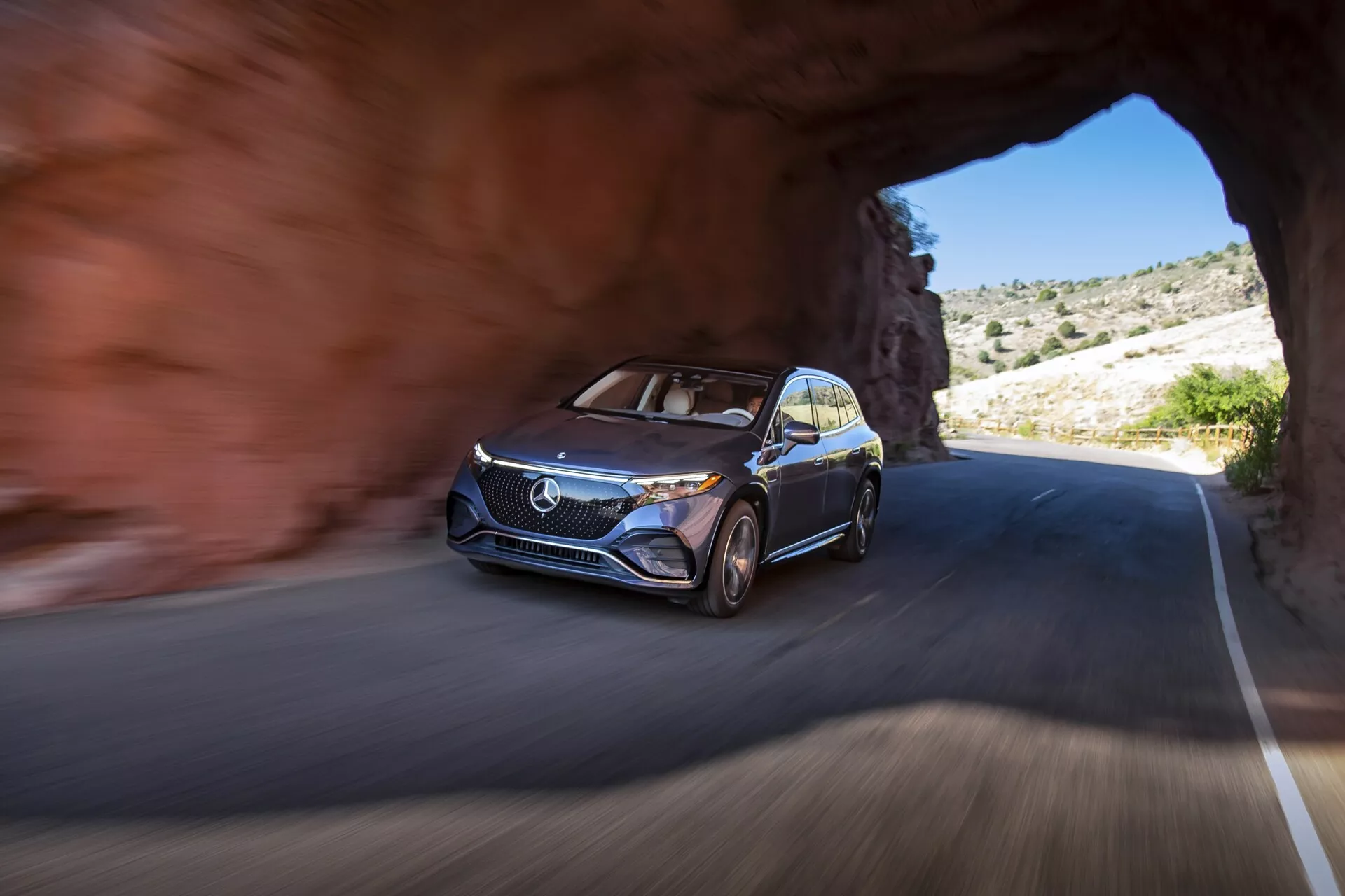Image d'un Mercedes EQS-SUV 2022 émergeant d'un tunnel sur une route, capturée sous un angle bas se concentrant sur le côté avant gauche, mettant en valeur le design moderne du véhicule avec une calandre proéminente, le logo, les feux de jour à LED, les jantes en alliage à rayons multiples et une finition de peinture métallique. La voiture est en mouvement, comme indiqué par l'arrière-plan et les roues flous. L'environnement montre un terrain rocheux avec une végétation clairsemée, suggérant une région montagneuse ou désertique sous un ciel dégagé.