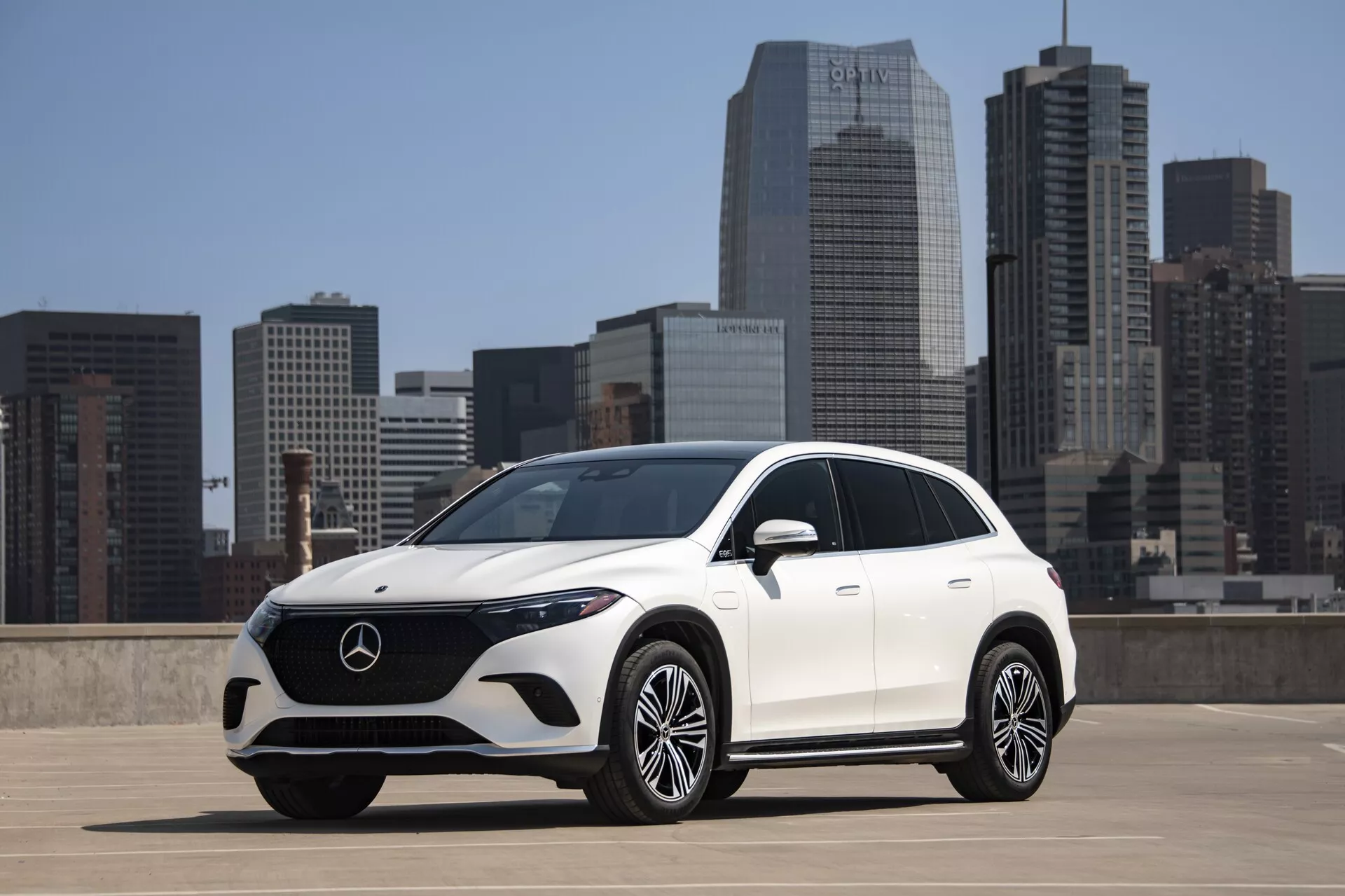 Vue avant trois quarts d'un SUV Mercedes-Benz EQS blanc de 2022 sur le toit d'un parking avec la ligne d'horizon de la ville en arrière-plan. Le SUV présente un design élégant avec des lignes lisses, une grande calandre avec l'emblème Mercedes-Benz et des phares LED. Le pare-chocs avant a de grandes prises d'air et une plaque de protection argentée, et les jantes en alliage à multiples rayons ajoutent une touche de luxe. La technologie avancée comprend des capteurs ou des caméras possibles dans la calandre avant, des clignotants intégrés dans les rétroviseurs latéraux, et le design aérodynamique suggère une optimisation de l'efficacité énergétique. La lumière du soleil projette des ombres vers la caméra, mettant en valeur le design du véhicule.