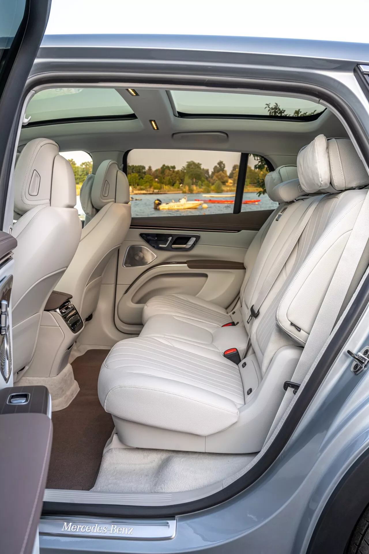 Vue intérieure du Mercedes EQS SUV 2022 depuis la porte passager arrière droite, mettant en valeur une banquette arrière en cuir crème clair de luxe avec trois appuie-têtes, un toit panoramique, des commandes électroniques, des bouches d'aération climatiques individuelles, des accents chromés, une grille de haut-parleur, une poche de rangement et un tapis de haute qualité.