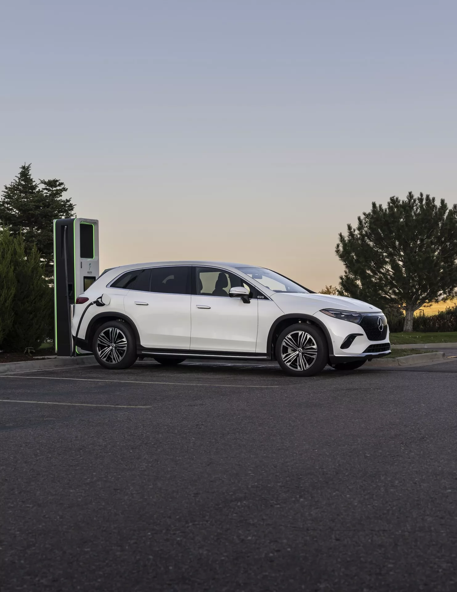 Moderne Mercedes EQS-SUV 2022 à une borne de recharge au crépuscule, photographié sous un angle latéral-frontal mettant en valeur son profil élégant, ses jantes en alliage et son port de recharge.