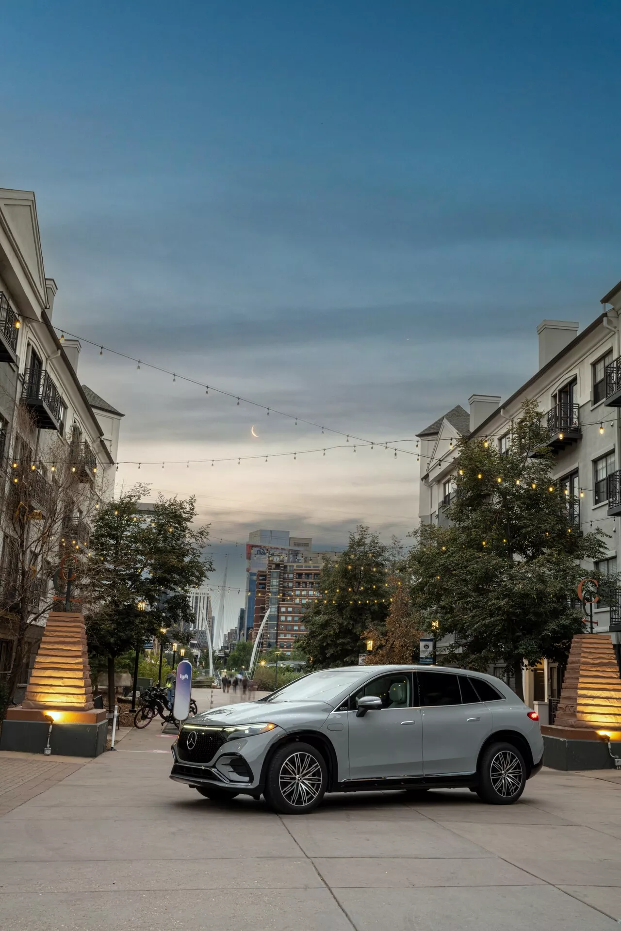 Vue à trois quarts de face d'une Mercedes EQS-SUV 2022 argentée mettant en avant sa calandre proéminente avec emblème, ses feux de jour à LED, ses jantes en alliage et son style de carrosserie. La voiture est stationnée dans un environnement urbain chic avec un éclairage décoratif et des bâtiments, mettant en avant son design luxueux et moderne.