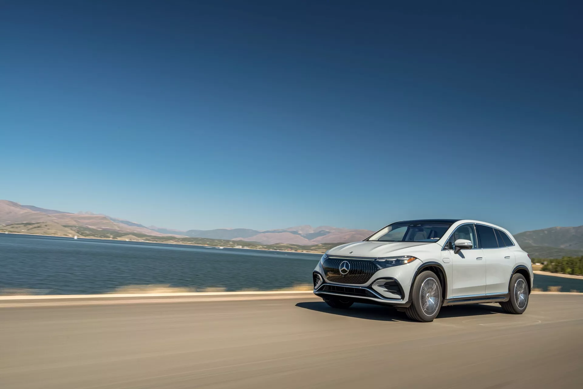 SUV Crossover de luxe moderne, Mercedes-Benz EQS, roulant sur une route goudronnée près d'un grand plan d'eau. L'image capture la voiture sous un angle frontal trois quarts en plein jour, mettant en évidence des caractéristiques telles que les phares à LED, une calandre distinctive, un design aérodynamique et de grandes jantes en alliage. Le véhicule est en mouvement, suggérant du dynamisme, avec un lac ou une rivière en arrière-plan et un ciel dégagé, emblématique d'un modèle électrique haut de gamme.