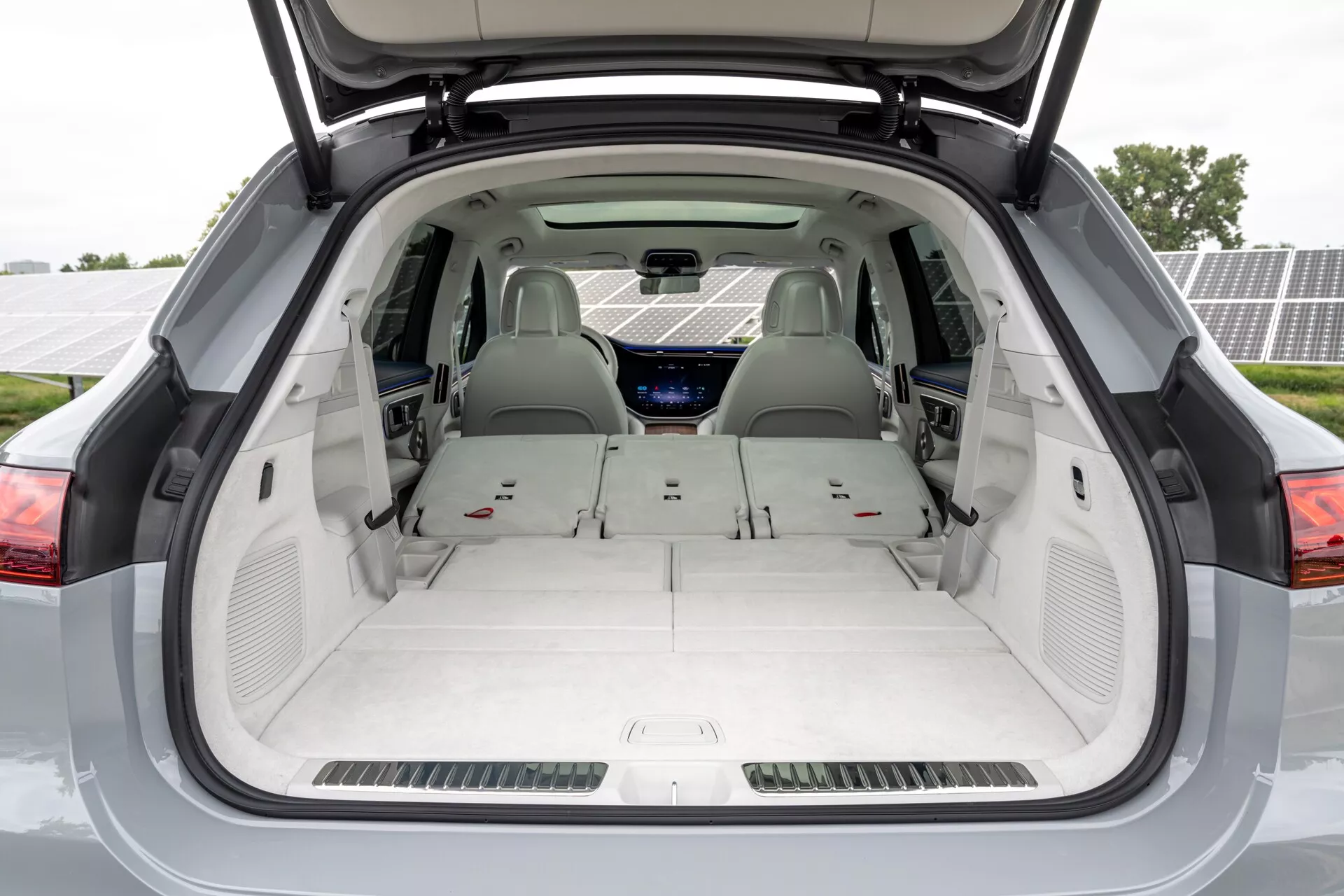 Vue arrière d'une Mercedes EQS-SUV 2022 depuis la zone de chargement avec le coffre ouvert, montrant un espace de chargement spacieux et plat avec les sièges arrière rabattus, une sellerie de couleur claire, un panneau de commande de climatisation arrière et un écran numérique visible sur le tableau de bord, indiquant peut-être un modèle électrique ou hybride de luxe à trois rangées, avec des panneaux solaires légèrement visibles en arrière-plan.