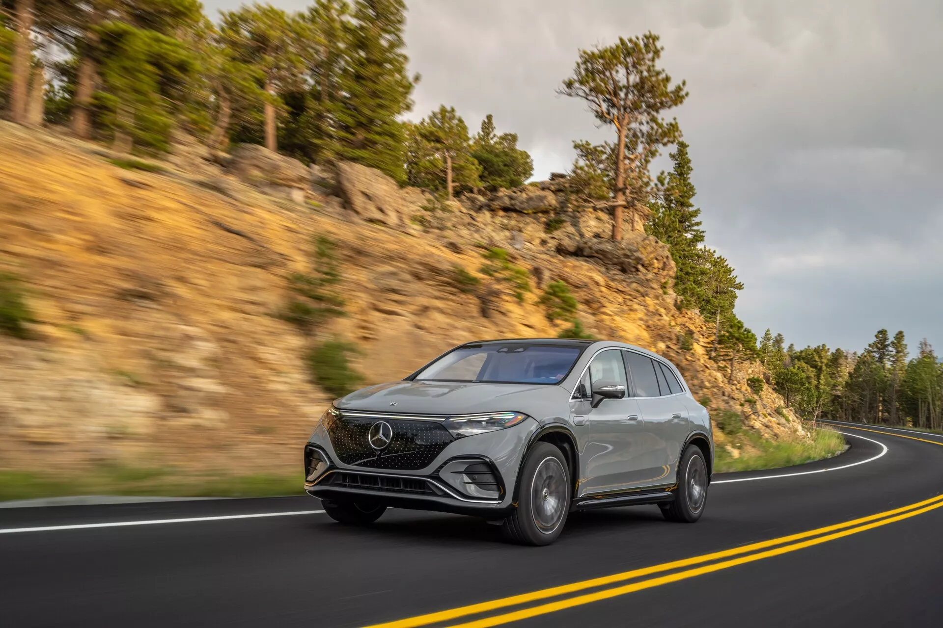 Prise de vue dynamique d'un Mercedes EQS SUV 2022 argenté sur une route de montagne sinueuse, vue sous un angle avant trois quarts pour accentuer sa hauteur et sa présence sur la route. Équipé de phares LED élégants, d'un badge frontal distinctif, de jantes en alliage de luxe et de lignes aérodynamiques indiquant un design contemporain.