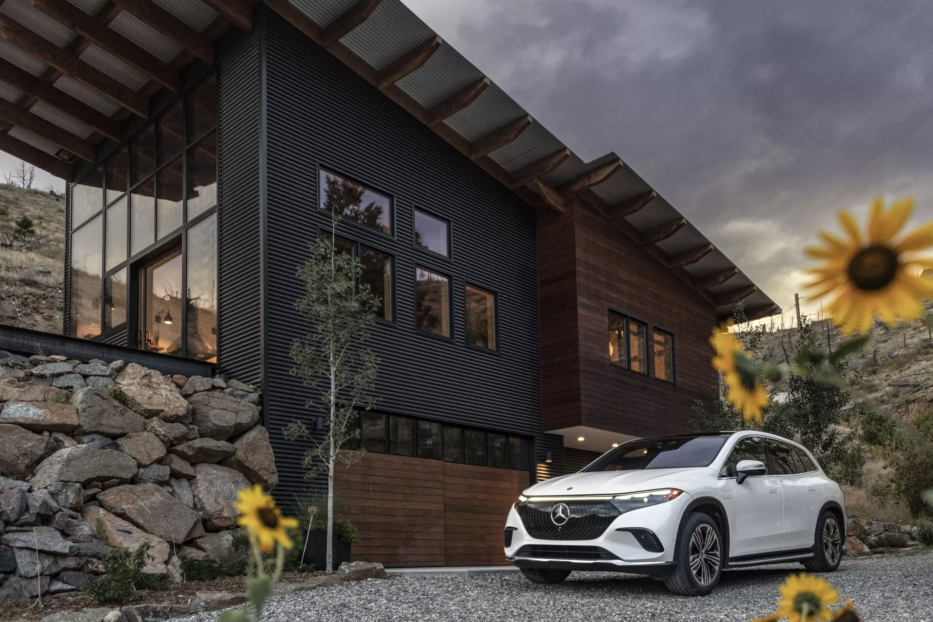 Image d'un SUV de luxe Mercedes-Benz EQS 2022 garé devant une maison contemporaine avec un ciel nuageux en arrière-plan. La prise de vue en contre-plongée met en valeur la posture et la taille du véhicule, en se concentrant sur la calandre avant, l'ensemble de phares, le pare-chocs avant, les contours du capot et le côté conducteur s'étendant jusqu'à la porte arrière. Les caractéristiques comprennent des feux diurnes à LED et des roues élégantes, ajoutant un sentiment de luxe et de technologie avancée. Le SUV blanc contraste avec la maison sombre et le paysage rocheux, entouré de fleurs légèrement floues en raison d'une faible profondeur de champ, mettant en avant le SUV comme point focal principal.