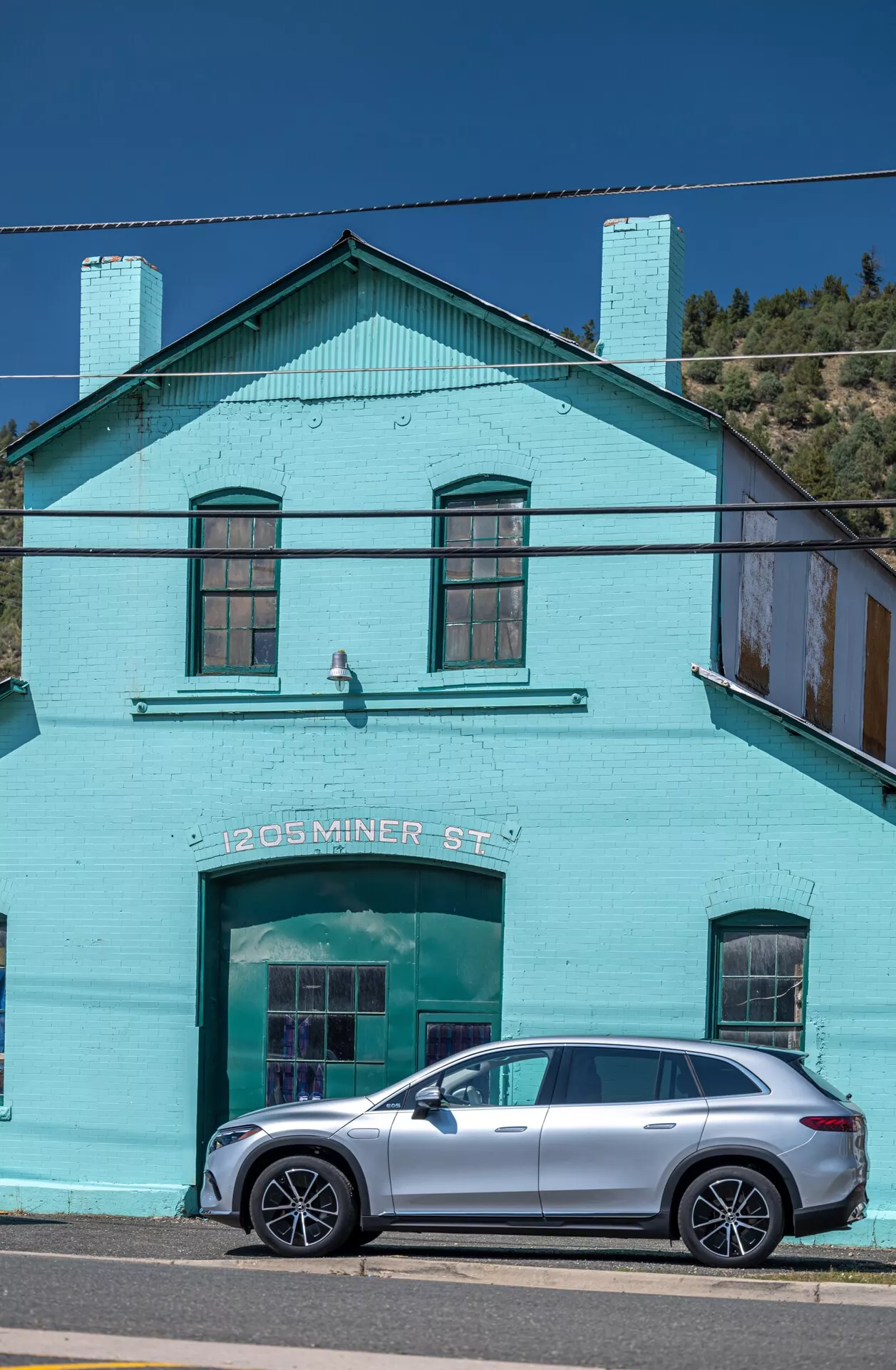 Vue de profil latéral d'un Mercedes EQS SUV 2022 argenté, garé sur le côté d'une rue devant un bâtiment à la façade turquoise distincte. Capturé sous un angle légèrement vers l'avant, la perspective à mi-hauteur montre le côté de la voiture, une partie de la calandre avant, les phares et une partie du capot. Le SUV présente un design élégant, des roues en alliage à cinq branches, des rétroviseurs avec clignotants intégrés, des barres de toit, le contour d'un toit ouvrant, des phares LED modernes, des poignées de porte de couleur carrosserie et des capteurs de stationnement sur le pare-chocs avant. La scène met en avant la vie quotidienne avec la voiture comme élément principal, sur le fond unique de '210 S Miner St.'