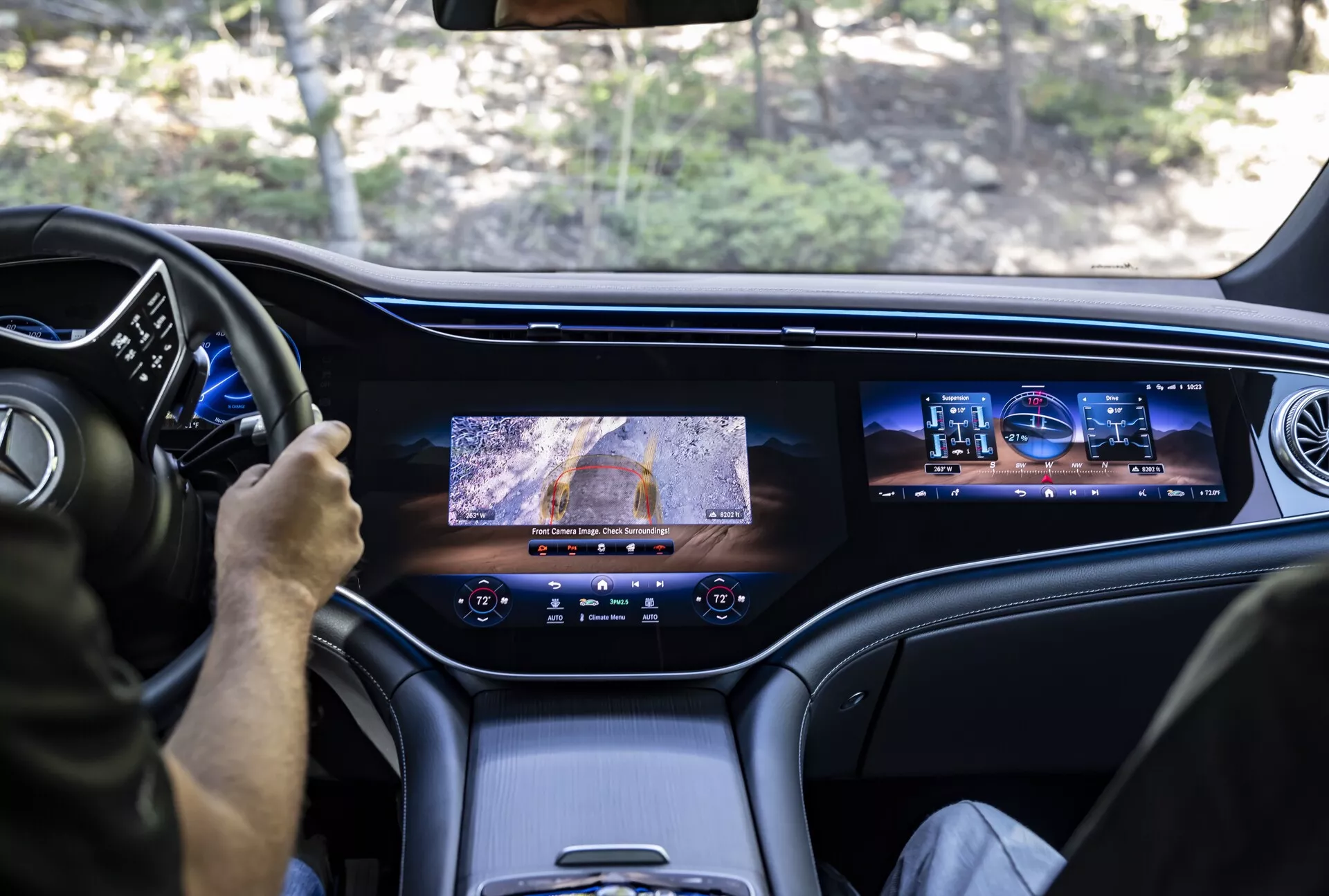 Vue depuis le siège arrière d'un Mercedes EQS-SUV 2022 montrant l'intérieur avant; affiche un conducteur utilisant le volant multifonction, le tableau de bord numérique, un grand écran central avec vue de caméra, un écran d'info-divertissement secondaire pour le passager, des matériaux haut de gamme ou de haute qualité dans l'habitacle et des bouches d'aération au design moderne, soulignant les caractéristiques de luxe et de haute technologie du véhicule.