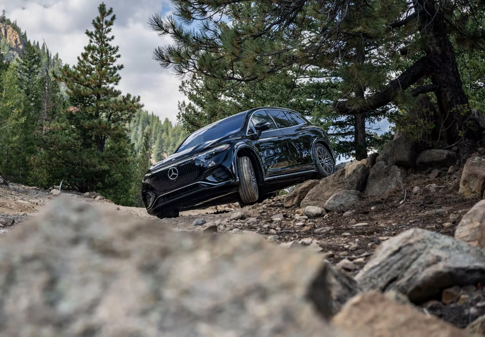 Prise de vue en contre-plongée d’un SUV Mercedes-Benz EQS noir de 2022 dans un cadre extérieur accidenté, montrant son côté avant gauche avec une vue partielle du côté droit. L’image met en évidence les phares distinctifs du véhicule, la grille avec l’emblème Mercedes, les roues en alliage et les pneus robustes sur une piste de terre avec des pierres en vrac, soulignant la capacité tout-terrain et le design de luxe.