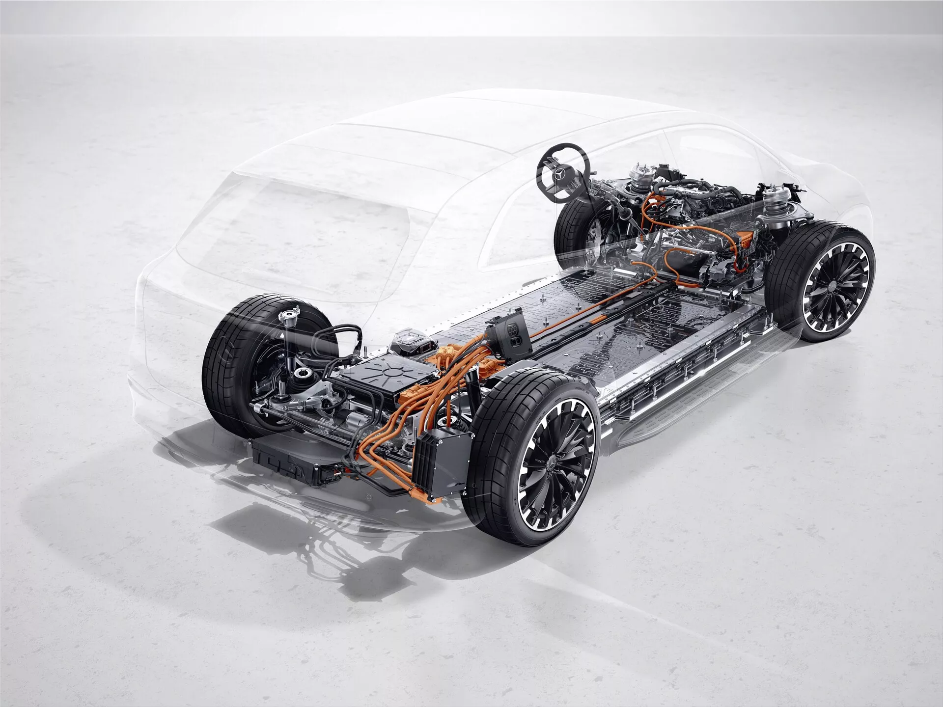 Photographie en studio du châssis transparent et du groupe motopropulseur du Mercedes EQS SUV 2022. L'image est prise sous un léger angle plongeant, mettant en avant le côté avant gauche. La photographie montre la structure interne, comprenant les essieux avant et arrière, les composants de la suspension, les roues et le pack de batteries placé sous le plancher, typique des véhicules électriques modernes. Les câbles orange visibles indiquent un système haute tension. Le groupe motopropulseur du véhicule, caractérisé par l'absence d'un moteur à combustion traditionnel, suggère la présence d'un ou plusieurs moteurs électriques. L'absence de système d'échappement et de réservoir de carburant confirme la nature électrique du véhicule. Le système de suspension indépendante indique des caractéristiques avancées de maniabilité et de qualité de conduite.