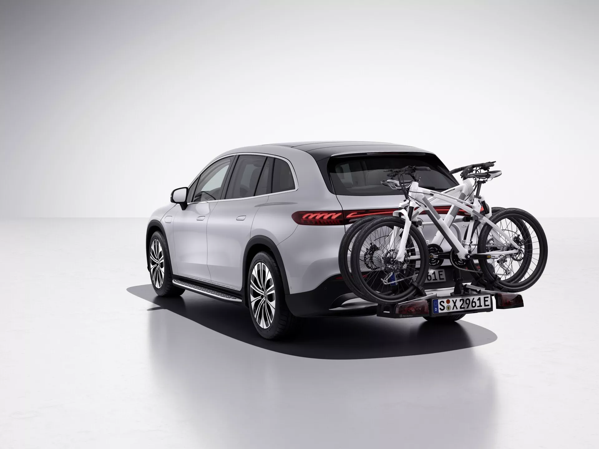 L'image présente un SUV Mercedes EQS 2022 dans un cadre de studio avec un arrière-plan blanc éclatant, vu sous un angle arrière de trois quarts mettant en avant le côté passager. Le véhicule est équipé d'un porte-vélo fixé à l'attelage de remorque arrière, tenant un vélo. Le SUV affiche un design élégant et contemporain avec des lignes lisses, un groupe de feux arrière distinctif avec des feux arrière à LED rouges horizontales, des jantes en alliage à plusieurs rayons et une carrosserie peinte dans une couleur claire, possiblement argentée ou grise, contrastée par des garnitures noires et des habillages d'arches de roue. Les rétroviseurs latéraux, les rails de toit et les garnitures de fenêtre ont un ton plus sombre, avec un toit panoramique ajoutant une sensation premium. Cette image met en avant des caractéristiques comme l'éclairage LED, le design sportif des roues et un porte-vélo, la rendant adaptée aux catégories comme Véhicules, Technologie Automobile, Équipements pour l'Extérieur/les Sports et SUV de Luxe.