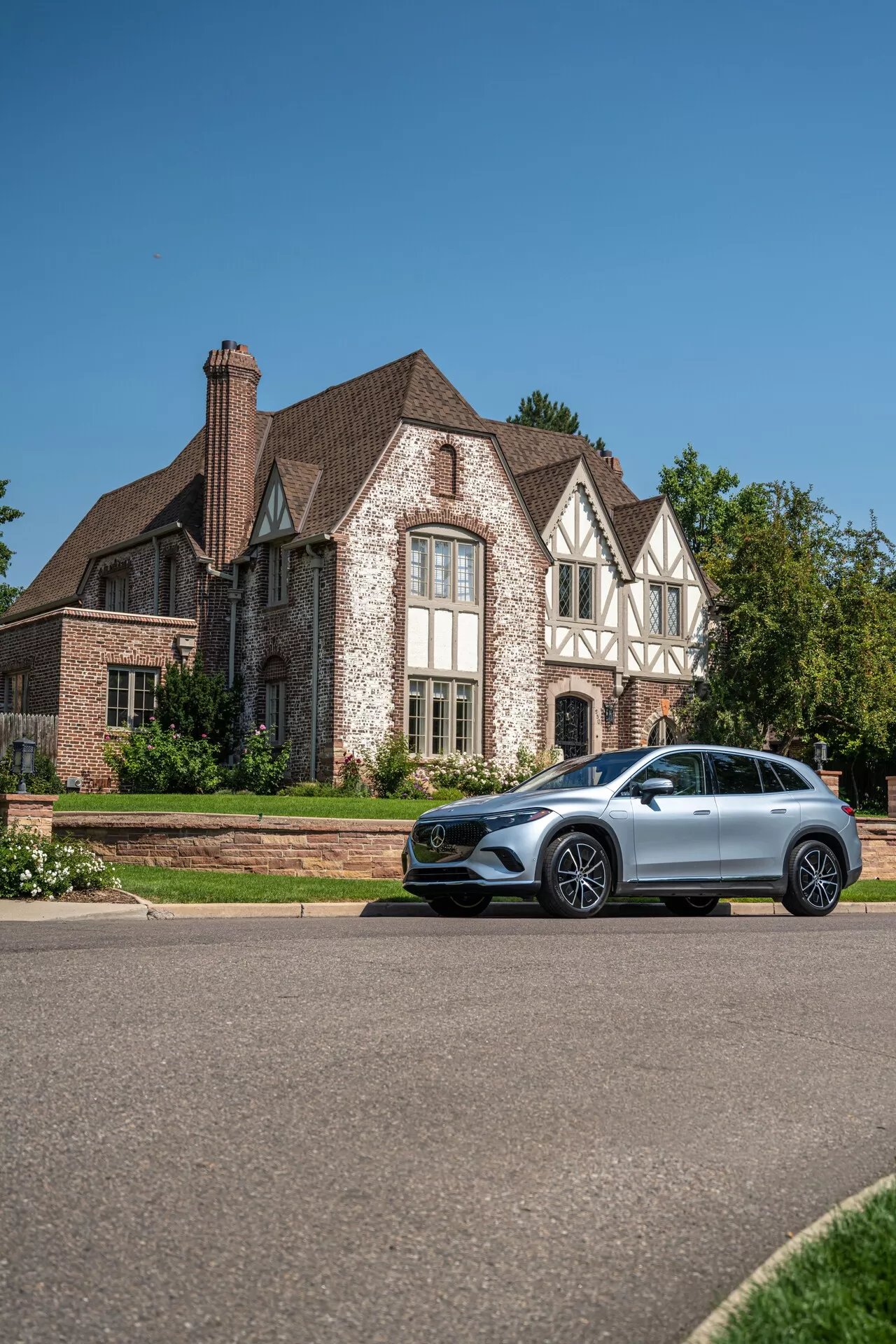 L'image montre un SUV Mercedes EQS 2022 garé sur le côté de la route devant une maison, prise d'un angle bas pour offrir une vue surélevée. Cet angle met en valeur le profil latéral et la face avant du véhicule, en soulignant des éléments de design tels que les grandes roues en alliage stylisées, les phares LED modernes, les rétroviseurs avec indicateurs de direction intégrés, le toit panoramique et les barres de toit. La voiture métallique, de couleur argentée ou grise et en excellent état, reflète son statut de luxe. L'environnement comprend une maison bien entretenue, renforçant l'impression d'opulence et de style de vie associée au véhicule.