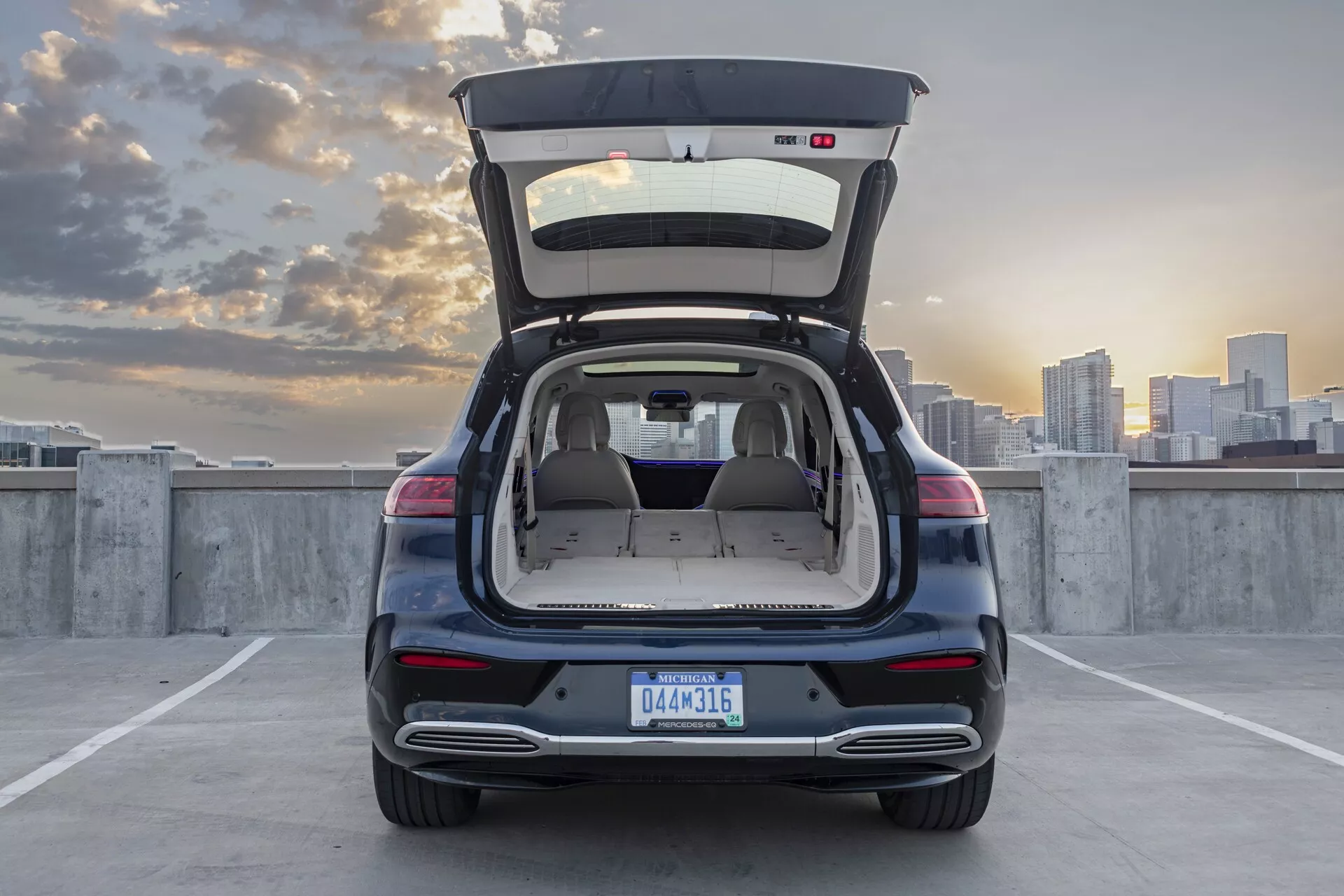 Vue arrière d'une Mercedes EQS-SUV 2022 photographiée sous un angle légèrement bas, mettant en avant l'espace du coffre ouvert. La voiture est garée sur le pont supérieur d'une structure de parking avec un ciel dégagé et des immeubles visibles au loin. La photo capture toute la largeur arrière, y compris les feux arrière, le pare-chocs arrière et une partie des roues arrière. L'éclairage ambiant et le ciel aux teintes pastel suggèrent que l'image a été prise au début de la soirée ou au crépuscule. L'image met en avant la zone de chargement du véhicule, le design des feux arrière, les réflecteurs et l'ouverture du coffre, s'appuyant sur la lumière naturelle pour des tons chauds et des ombres douces.